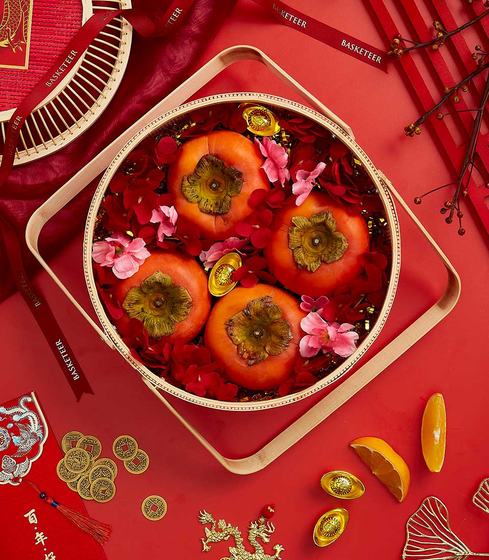 A top-view of a festive arrangement featuring four persimmons in a round bamboo basket, surrounded by red and pink flower petals. The scene is set against a red background with Chinese New Year decorations including gold ingots, coins, and red envelopes.