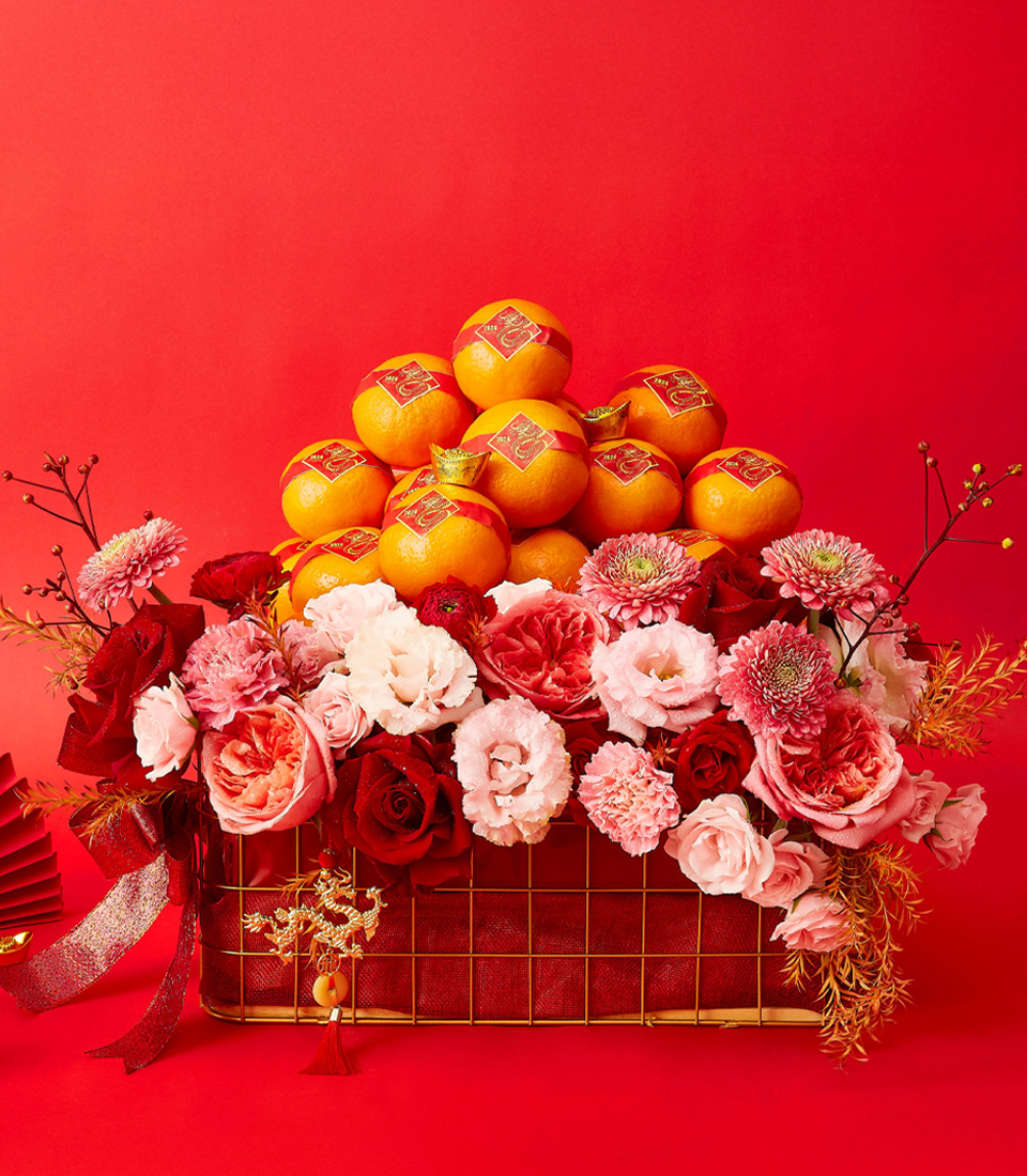 A decorative arrangement features a pyramid of oranges with red and gold stickers on top, set against a vibrant red background. Below the oranges, there is a lush assortment of pink, red, and white flowers, creating a colorful and festive display.