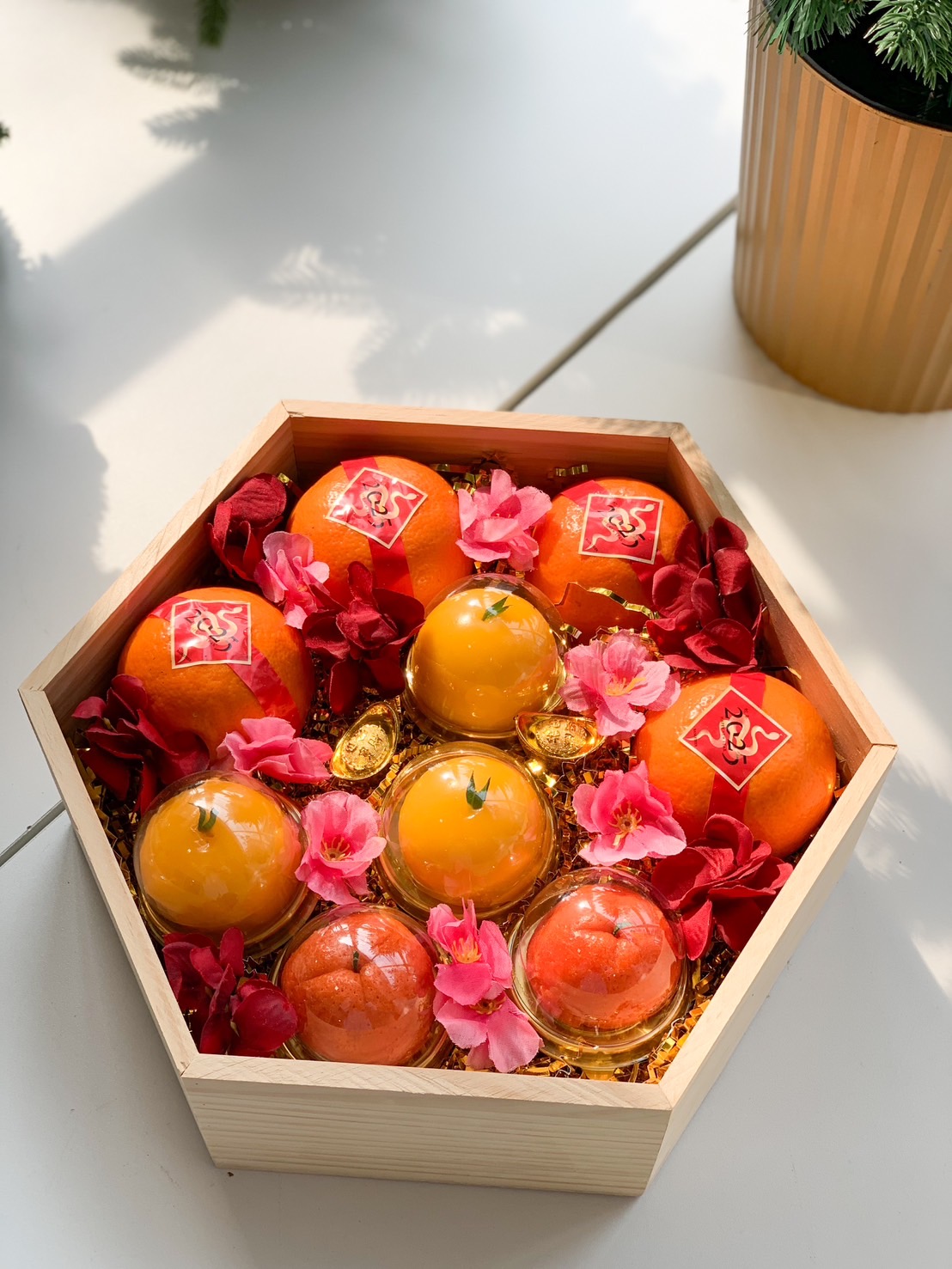 A festive hexagonal wooden gift box containing oranges, gold-wrapped spheres, and decorative flowers. Surrounding the box are gold ingots, artificial red foliage, a red pocket, and gold coins, all arranged on a red background for a celebratory occasion.