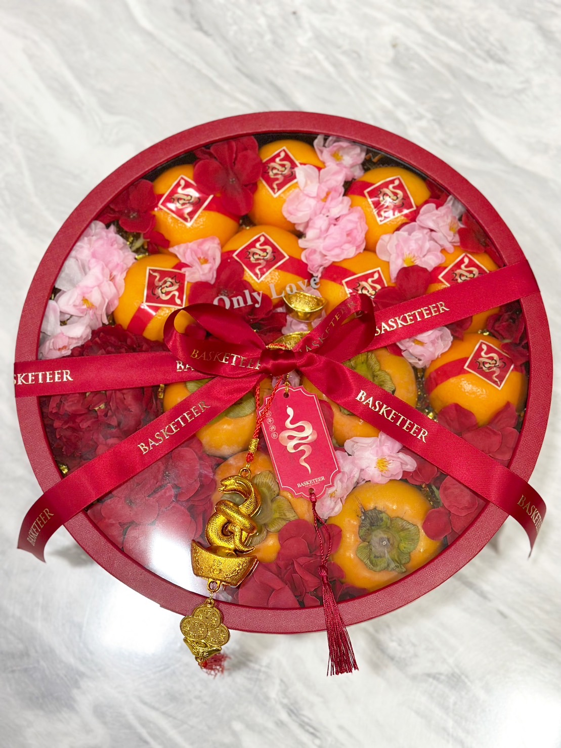 Round Chinese New Year gift box with fresh persimmons, floral decorations, golden charms, and red ribbon.