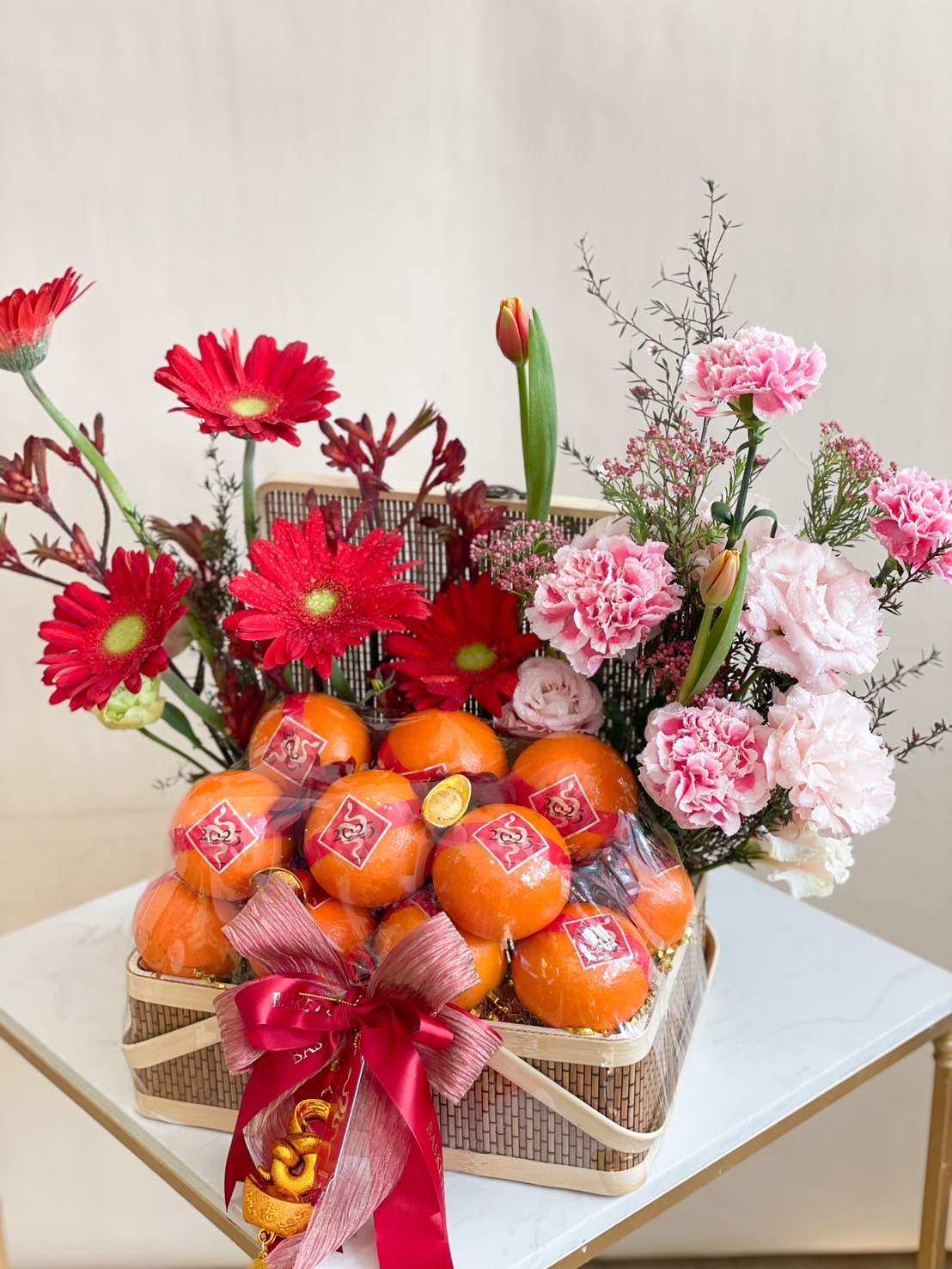 A decorative basket filled with vibrant pink and red flowers, including gerbera daisies, carnations, and lilies, alongside a bundle of mandarin oranges wrapped in a net. The basket is tied with red ribbon and placed on a white surface against a plain background.