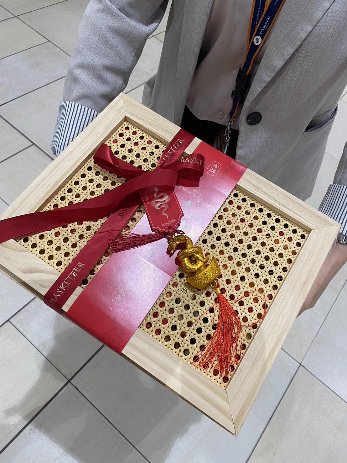 A stylish Chinese New Year gift box featuring a rattan weave design, red decorative ribbon, and a golden ornament with tassels, held by a person in a light gray blazer and white shirt, standing indoors on a tiled floor.