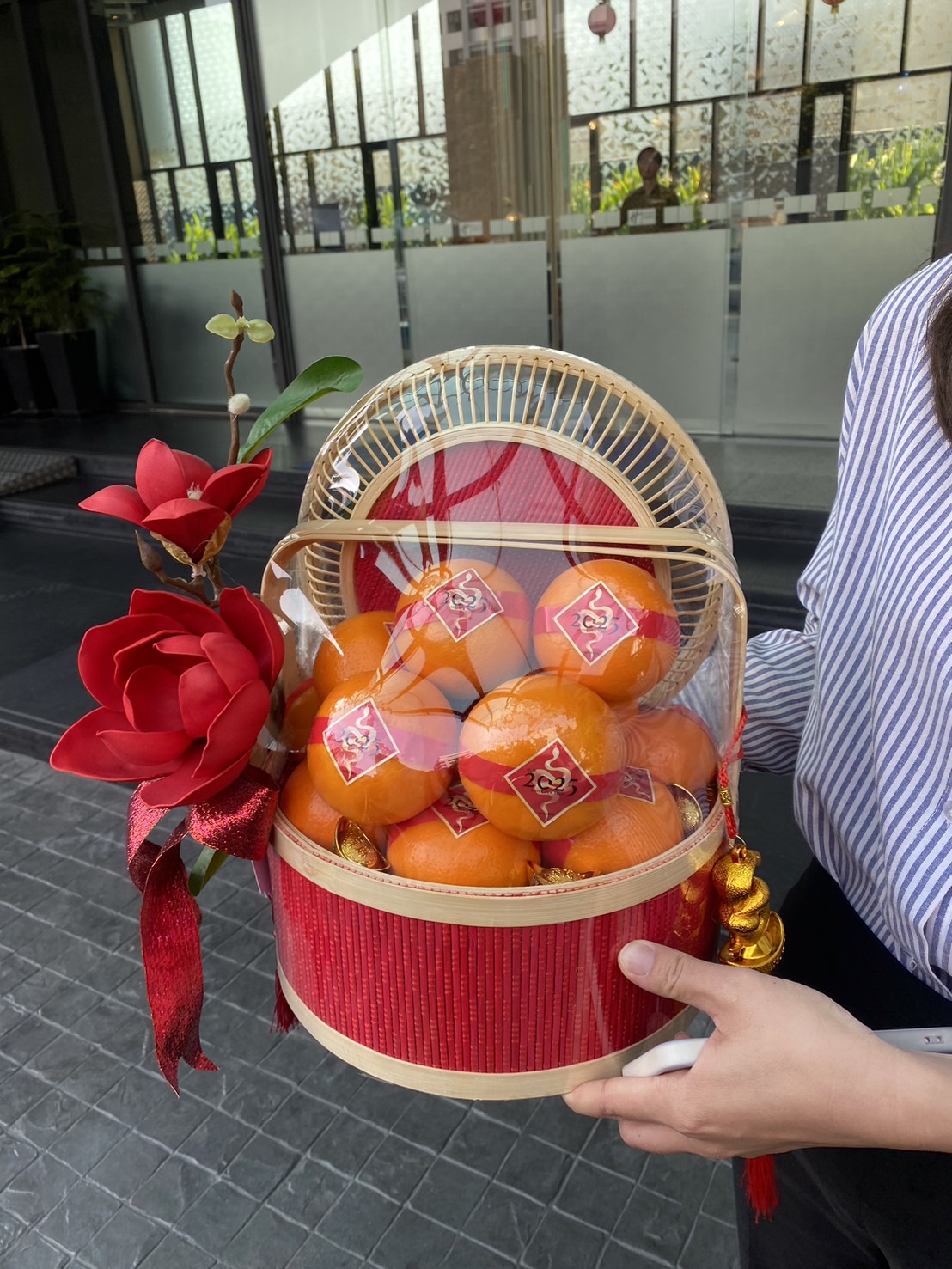A person holding a CNY orange gift basket filled with oranges and wrapped with a red ribbon stands near the entrance gate of a house. The gate is partially open, revealing a stone pillar and some greenery in the background.