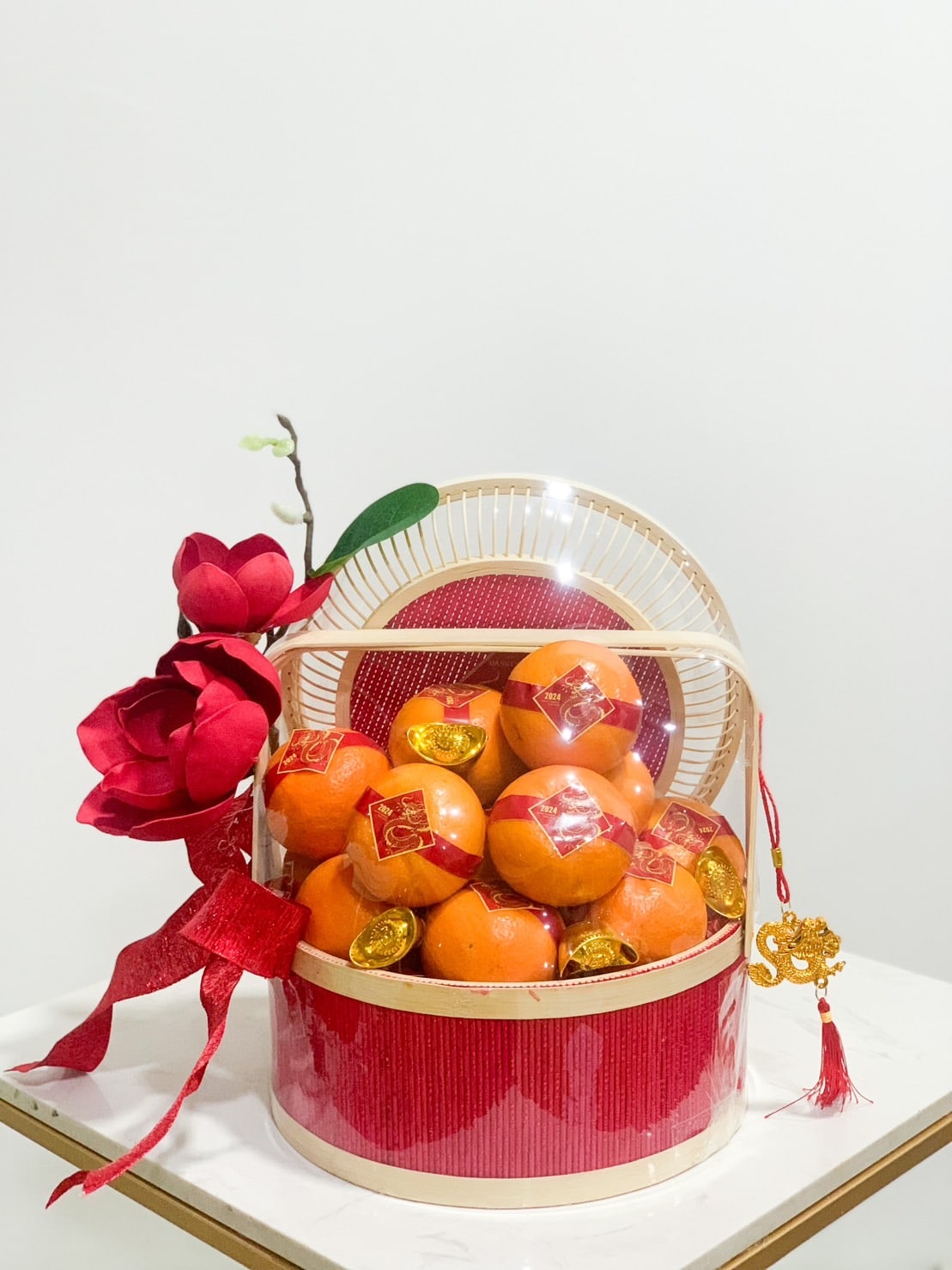 A CNY-themed orange gift basket filled with oranges wrapped in red squares, accompanied by gold coins. The basket is adorned with red fabric bows and a large red flower on the left, with a small branch extending upward. It rests on a white surface against a plain background.