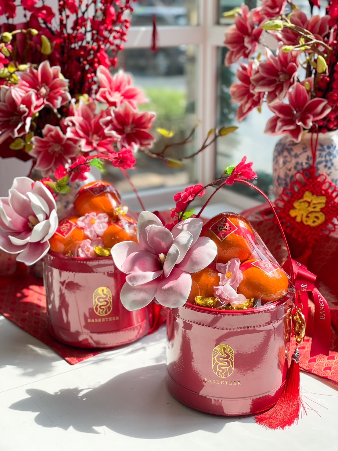 A beautifully designed Chinese New Year gift set featuring fresh mandarin oranges, gold-wrapped chocolates, and festive floral decorations in a pink luxury box.