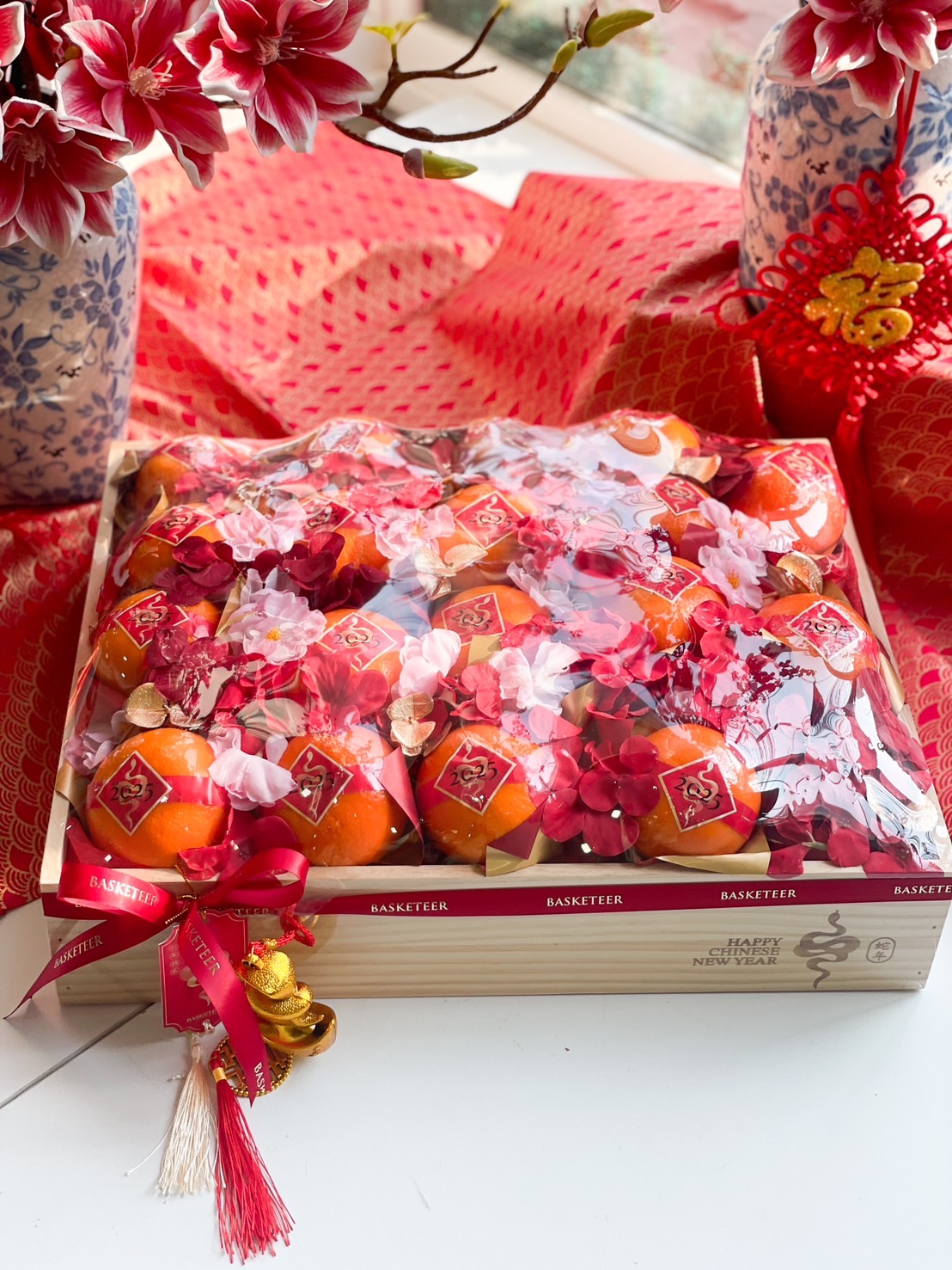 Chinese New Year wooden fruit box with fresh Mandarin oranges, red floral accents, and gold prosperity charms for a festive and elegant celebration.