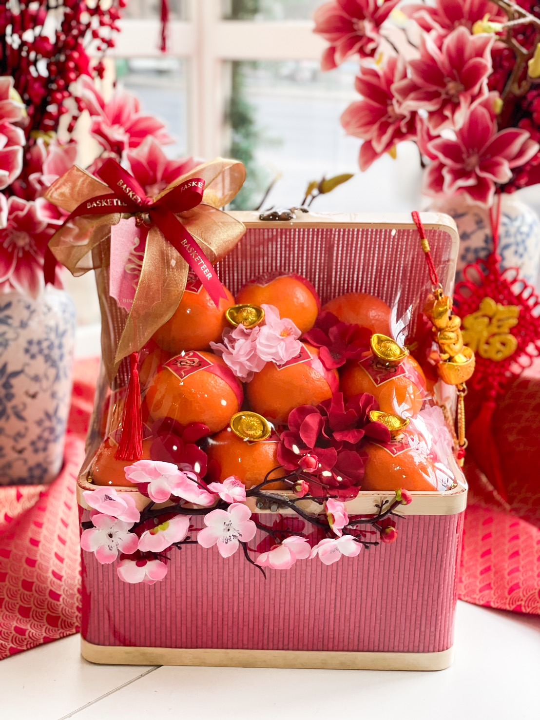 Elegant Chinese New Year gift box filled with fresh mandarin oranges, adorned with cherry blossoms, golden charms, and a festive red and gold ribbon.