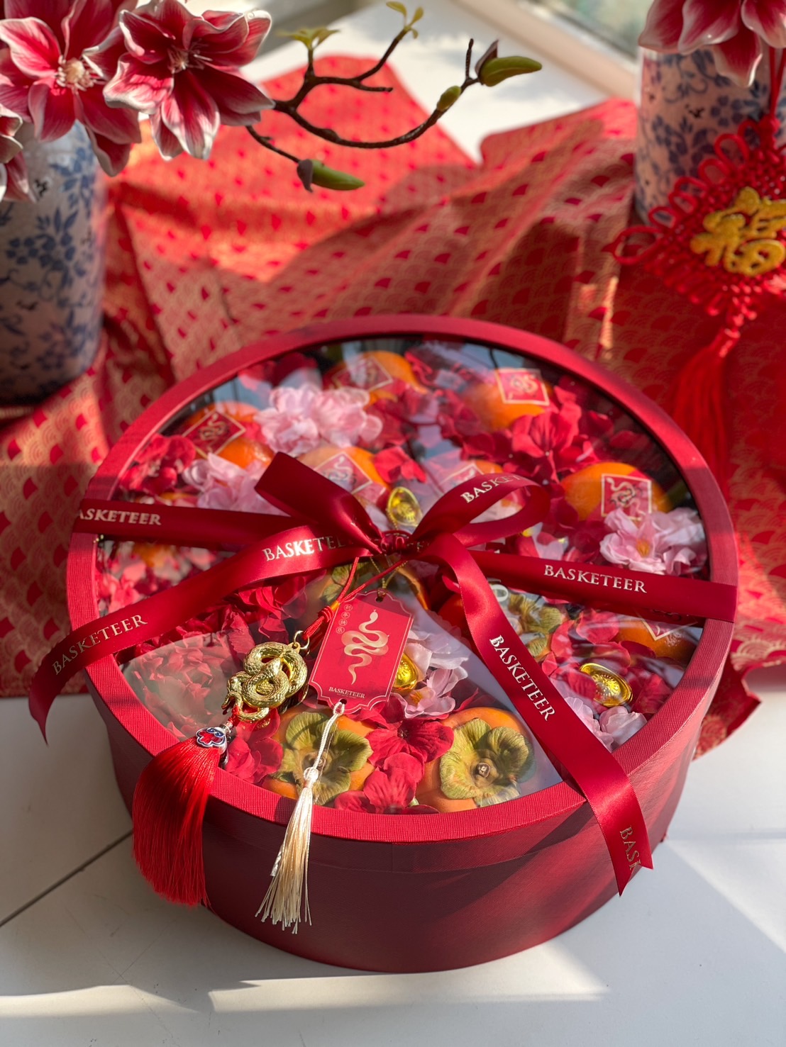 Luxury Chinese New Year round gift box with Mandarin oranges, festive flowers, and elegant red and gold decorations for prosperity and luck.