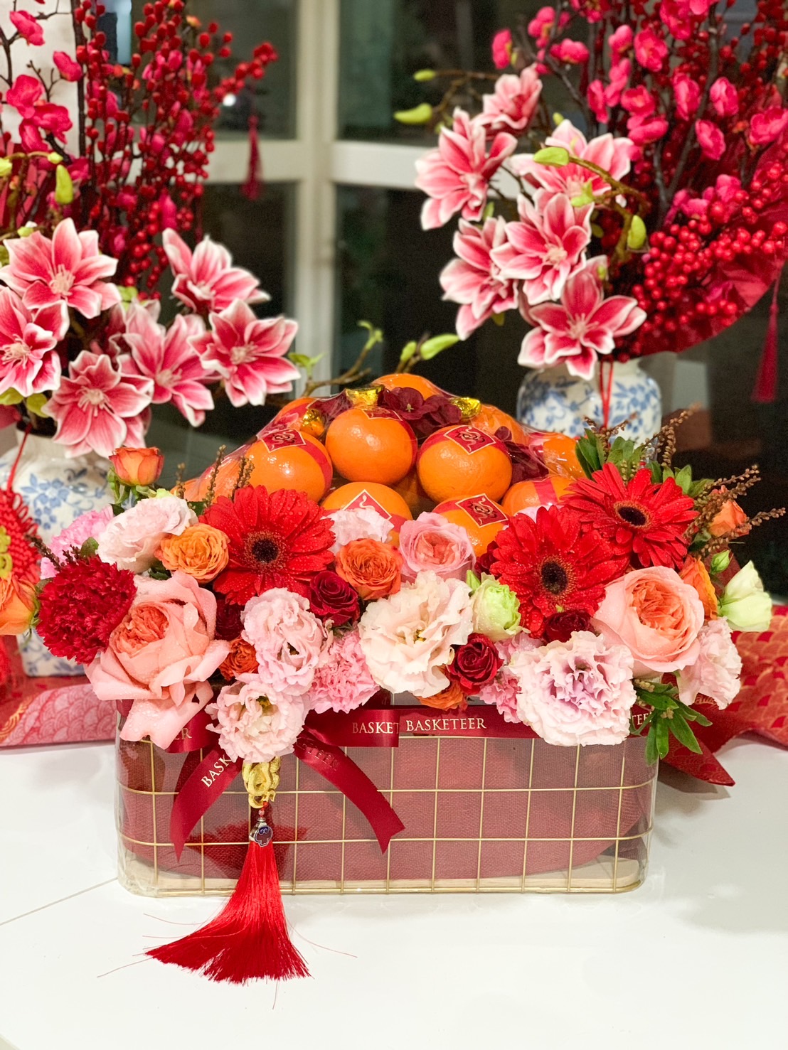 Chinese New Year gift basket with premium Mandarin oranges, vibrant red and pink flowers, and festive decorations for a prosperous celebration.