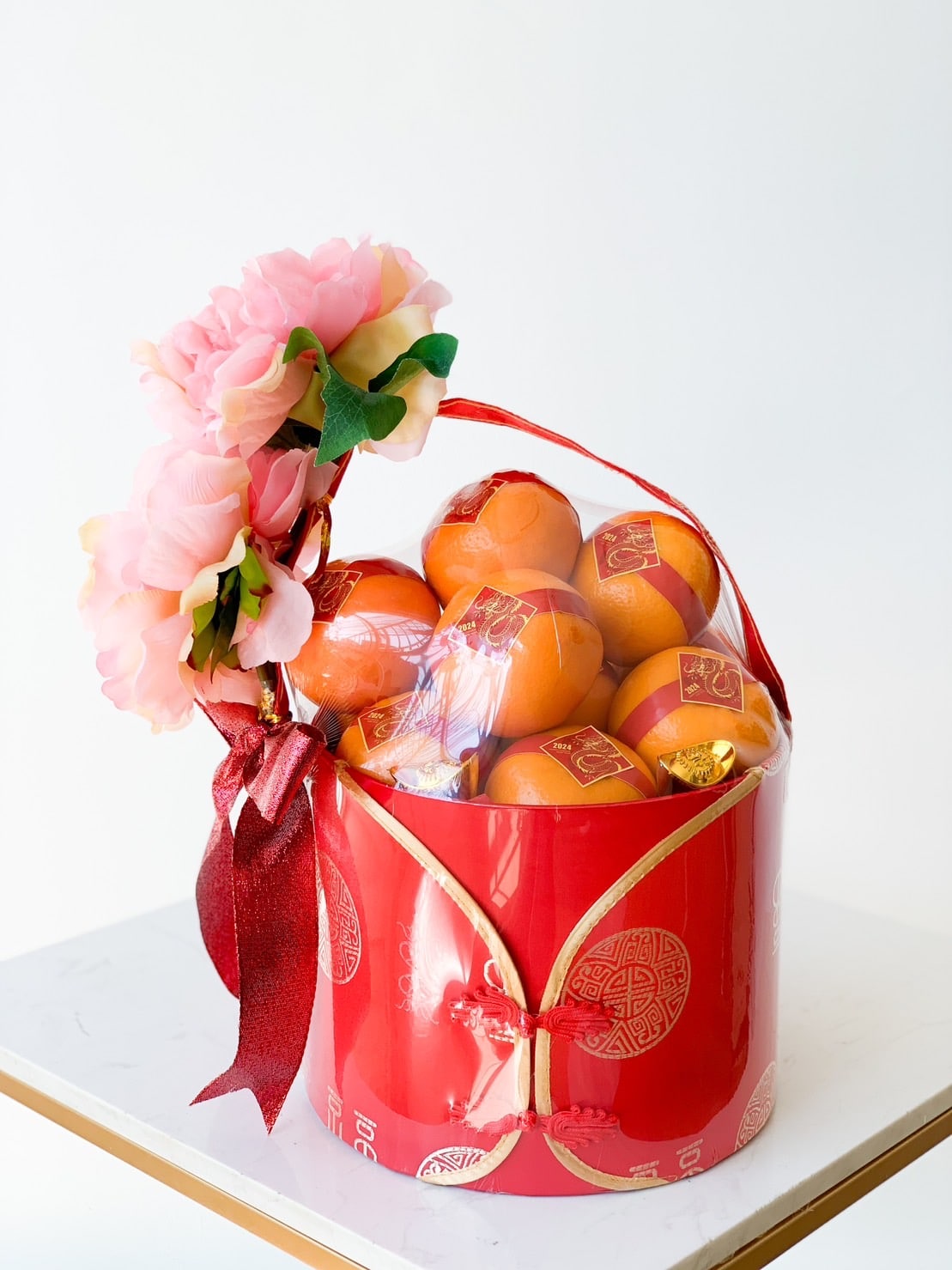 A festive red basket adorned with pink flowers and a red ribbon, filled with oranges wrapped in plastic. The basket features a traditional Chinese design, symbolizing good luck and prosperity. The arrangement is set against a plain white backdrop.