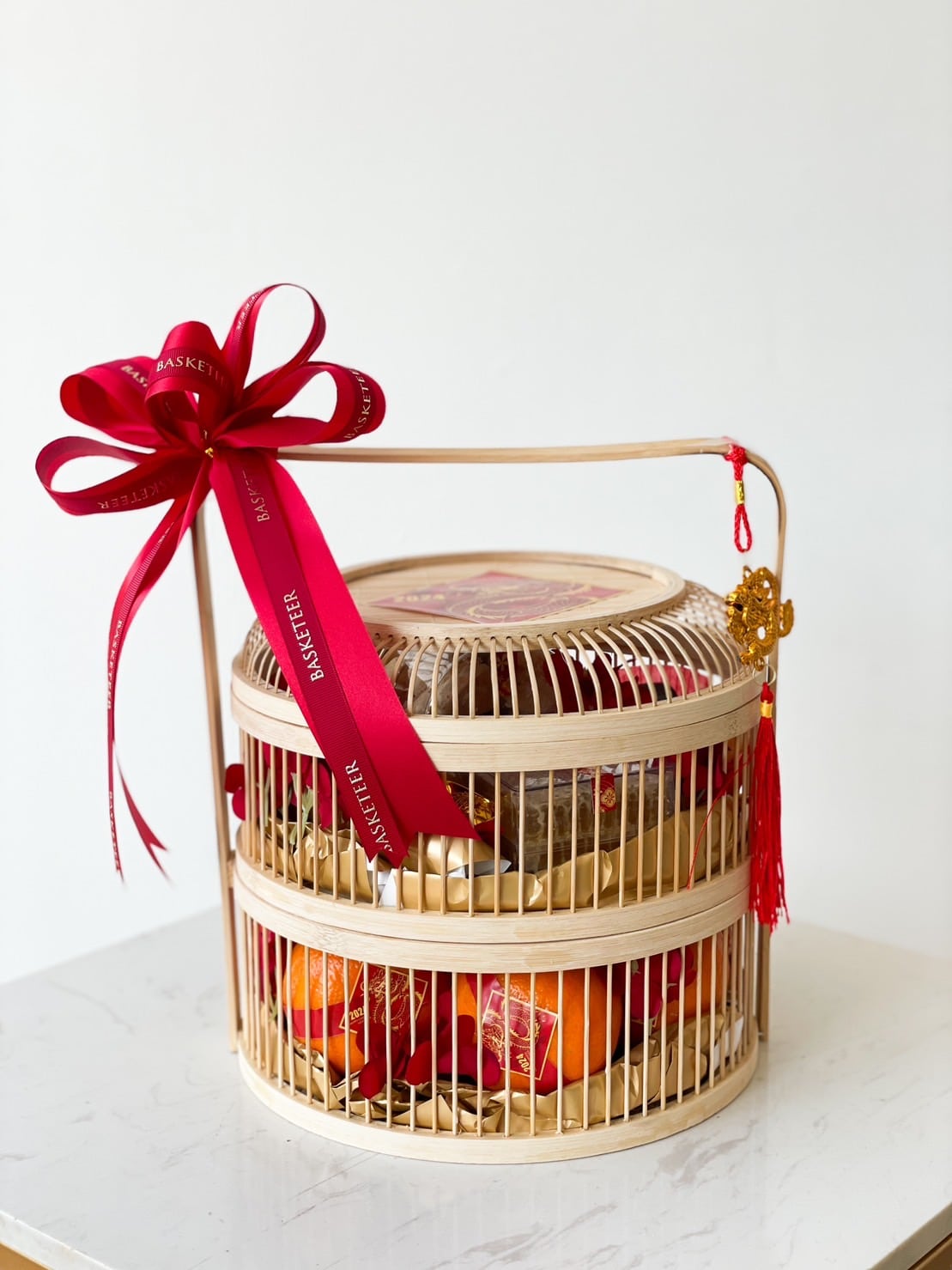 A two-tiered wicker basket adorned with a red ribbon and bow, holding various items including oranges and other goods. The basket is decorated with a small red tassel and golden ornament, resting on a white surface with a plain background.