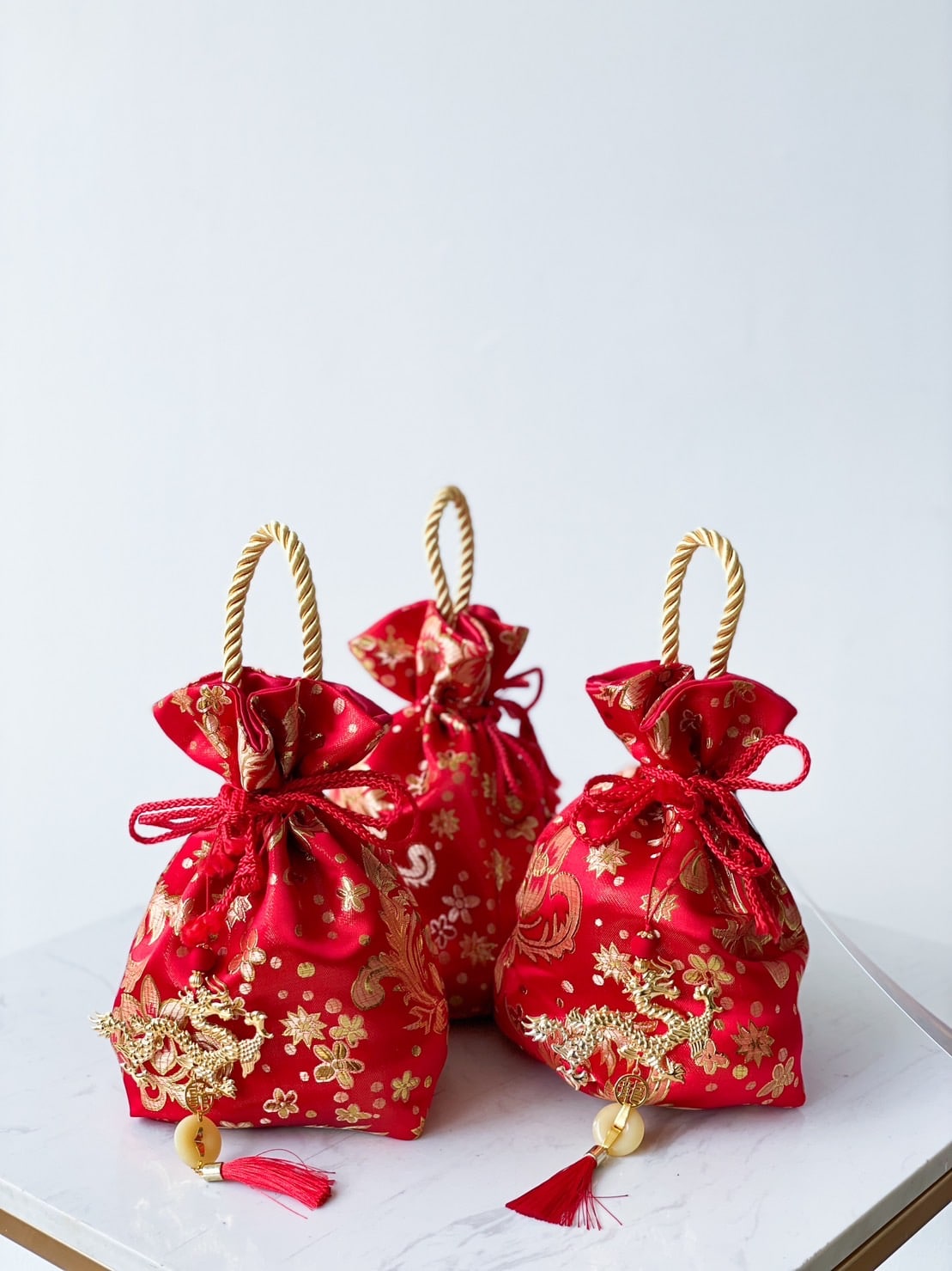 Three red drawstring gift bags with gold floral designs sit on a white square surface. These Mandarin Blessings Bags, perfect for CNY celebrations, have gold braided handles and are adorned with red tassels at the bottom. The background is plain white.