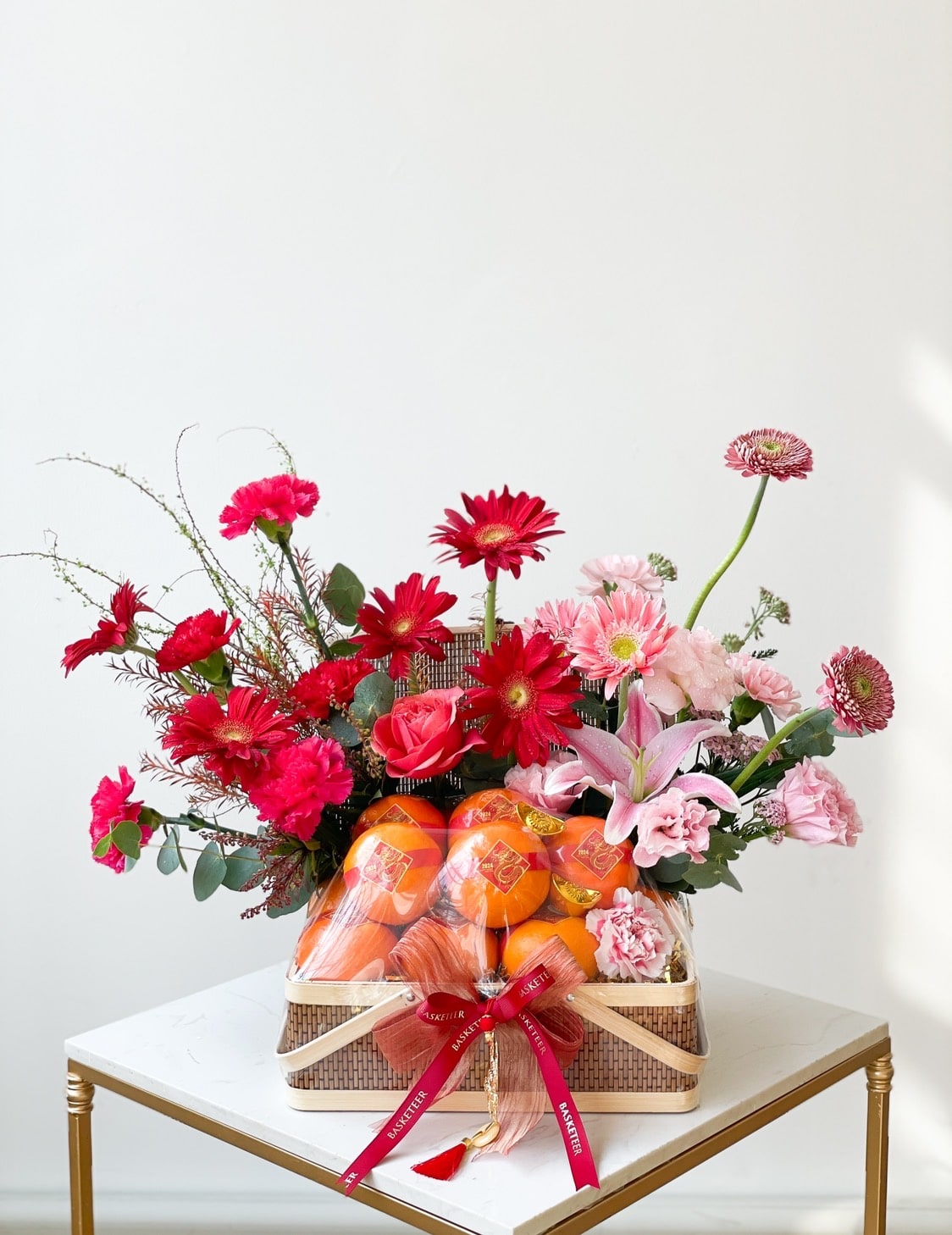 A vibrant floral arrangement featuring red and pink flowers, including roses, lilies, and daisies, displayed in a wicker basket. The basket also contains bright orange fruits wrapped with a red ribbon, all set on a small, square table against a white background.
