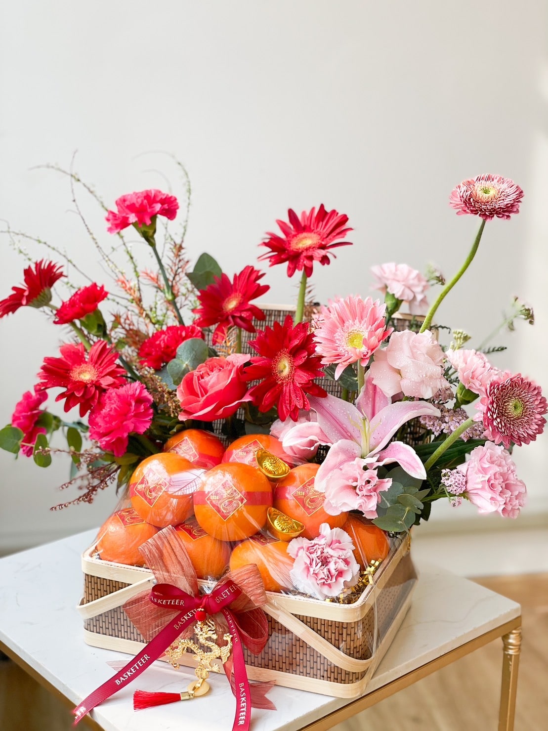A decorative basket filled with vibrant pink and red flowers, including gerbera daisies, carnations, and lilies, alongside a bundle of mandarin oranges wrapped in a net. The basket is tied with red ribbon and placed on a white surface against a plain background.