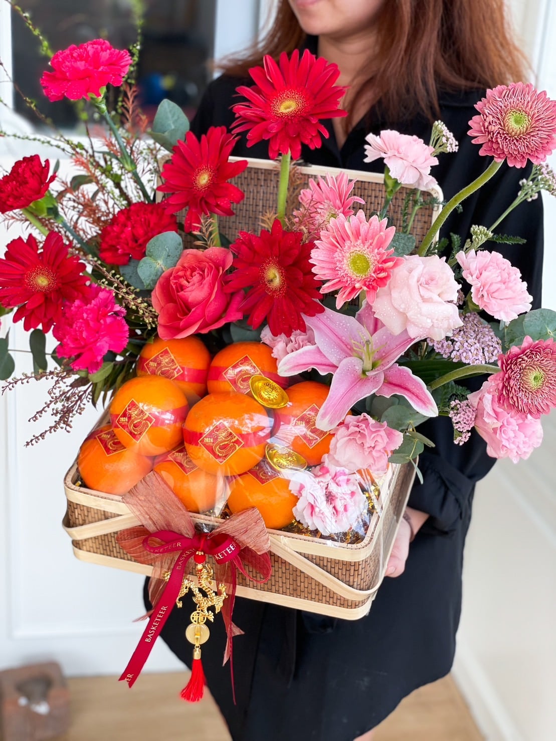 A person wearing a black outfit holds a decorative basket filled with bright red and pink flowers, including gerberas, roses, and lilies, alongside several persimmons wrapped in protective netting and packaging. The basket is adorned with a red ribbon and festive ornaments.
