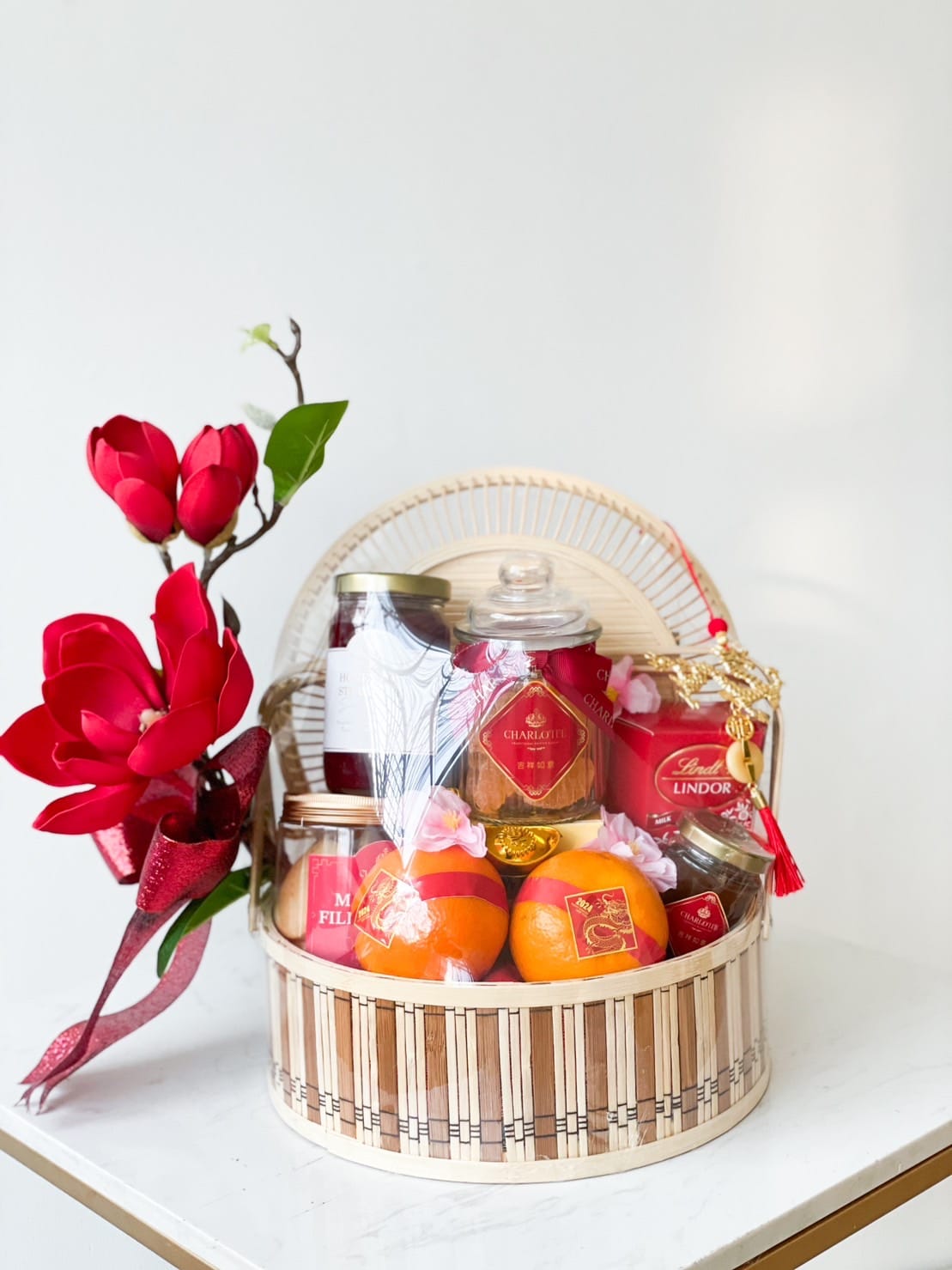 A festive gift basket containing various items like Lindor chocolates, jars, oranges, and a decorative glass jar. The basket has a natural woven texture, and a red magnolia flower arrangement adorns the left side, creating a vibrant and elegant display.
