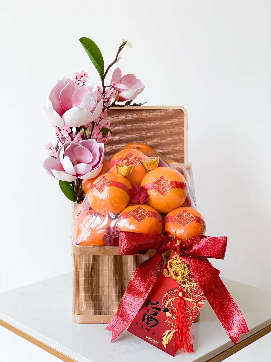 The Joyful Mandarin Bliss Hamper features a wicker basket filled with oranges, each wrapped in red and gold decorative paper. Adorned with magnolia flowers, tied with a large red bow, and including a red envelope with Chinese characters and a hanging ornament, it embodies the essence of CNY against a plain white background.