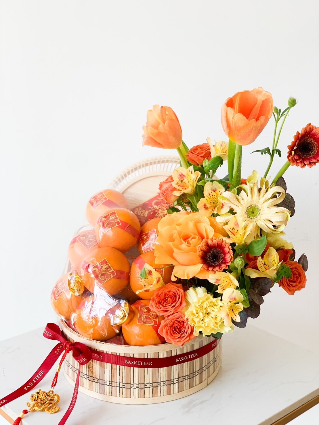 A decorative basket filled with bright orange persimmons wrapped in plastic sits on a table. The basket is adorned with a red ribbon and features a colorful bouquet of orange tulips, yellow daisies, and other vibrant flowers on one side.