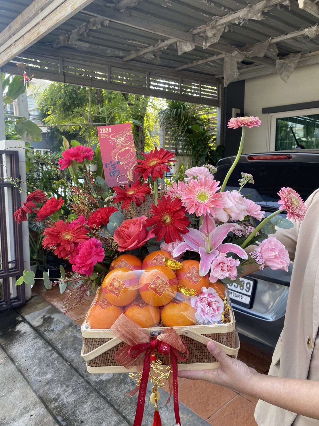 A person is holding a festive basket under a carport. The basket is filled with red and pink flowers, oranges, and decorative red envelopes. The basket is adorned with red ribbons and bows, with a car and greenery in the background.