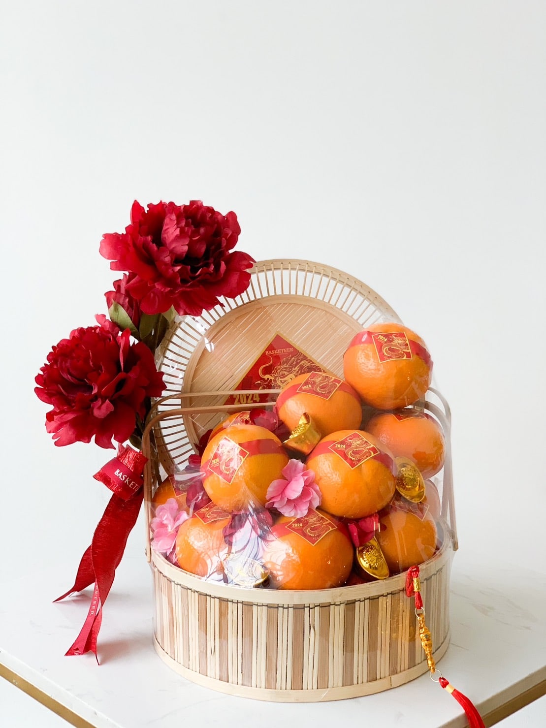 The Imperial Mandarin Elegance Basket, perfect for CNY festivities, features a decorative gift basket filled with oranges adorned with gold and red paper accents. It is accompanied by red flowers and red ribbons, all set against a plain white background.