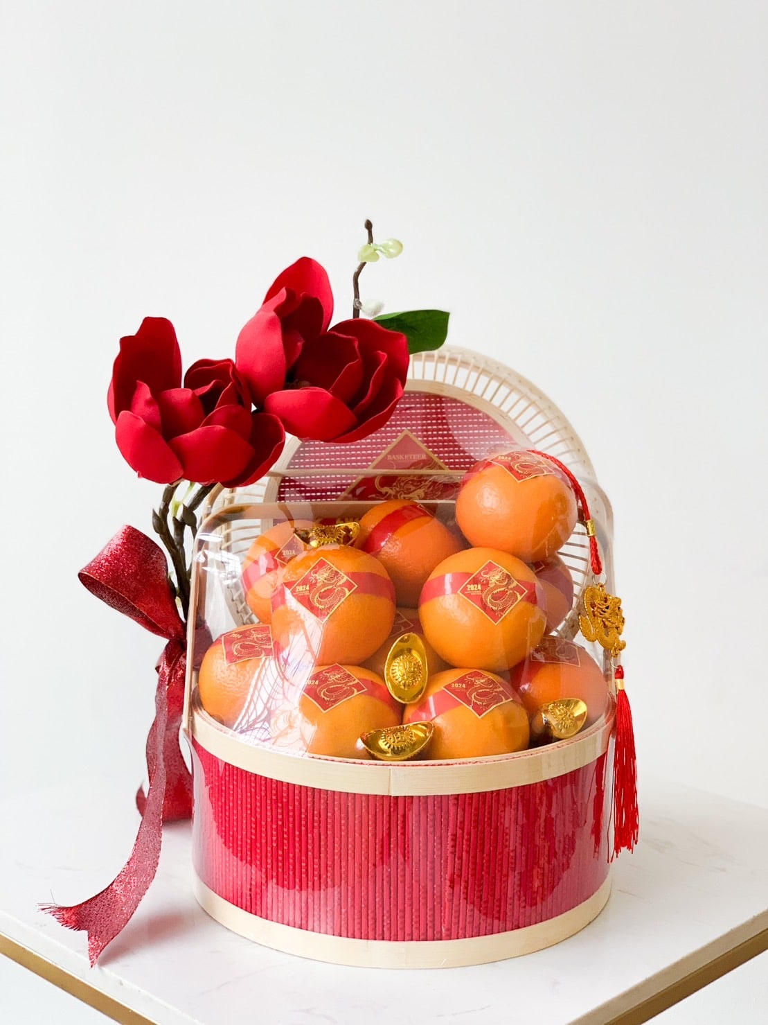 A decorative CNY Orange Gift Basket filled with oranges, adorned with red faux flowers, gold ingots, and a red ribbon. The basket has a white and red color scheme and a hanging red tassel with a golden charm, sitting on a white surface against a plain background.
