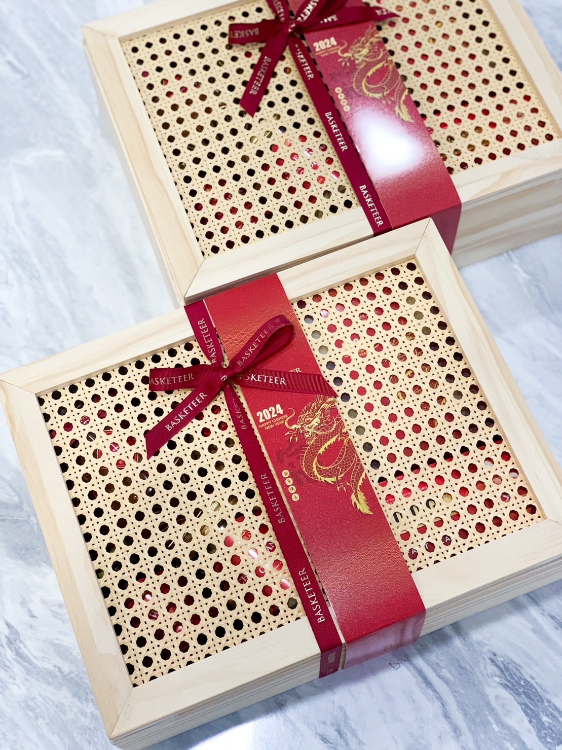 Two wooden gift boxes with intricate lattice tops are adorned with red ribbons. The boxes are arranged on a light-colored marble surface. The ribbons feature gold text and dragon designs, suggesting a festive or celebratory occasion, possibly related to the year 2024.