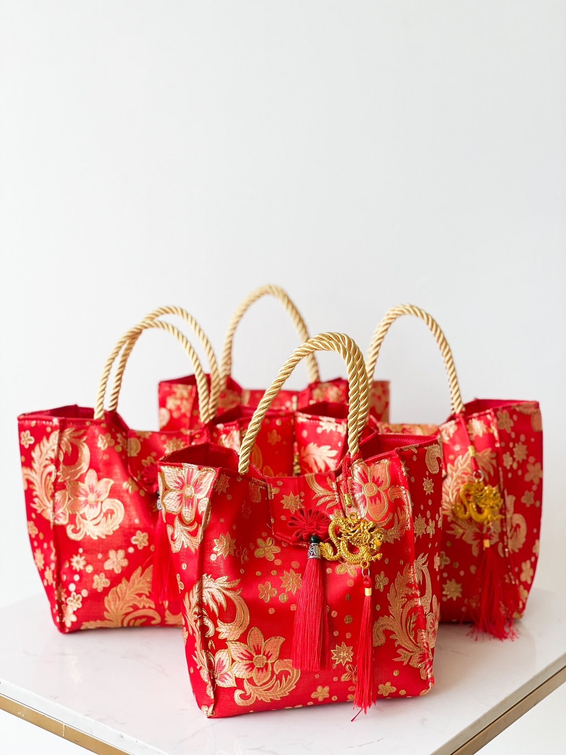 Three red and gold fabric bags with woven rope handles are displayed on a white surface. These CNY-themed citrus gift bags feature floral patterns and decorative tassels, elegantly arranged against a plain white background.