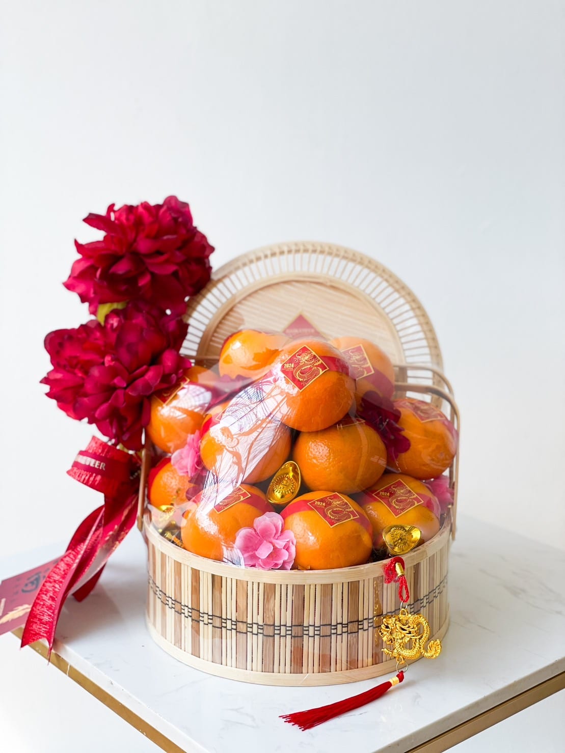 Behold the Imperial Mandarin Elegance Basket: a festive CNY basket filled with oranges, some wrapped in shiny red packaging, adorned with vibrant pink flowers. The basket is decorated with a big red flower and ribbons, and a small gold ornament hangs from it, all set on a pristine white surface.