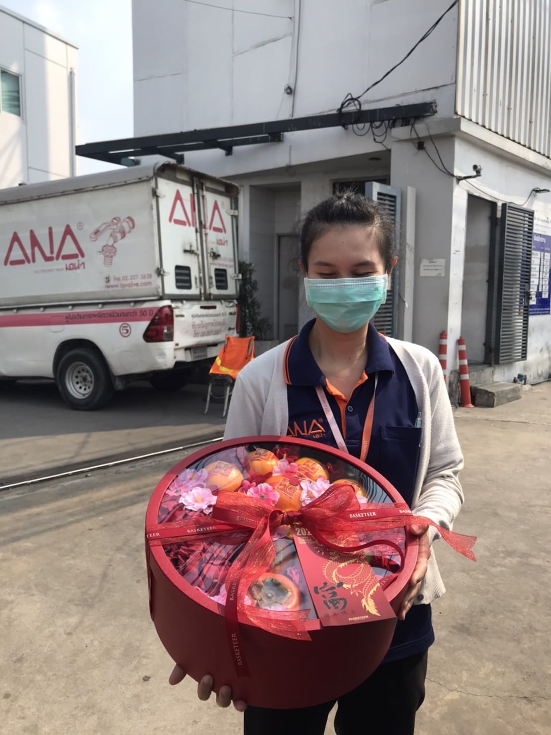 A woman wearing a face mask and a uniform stands outside a building, holding a large, round, red gift basket decorated with a ribbon. In the background, there is a white truck with the logo 