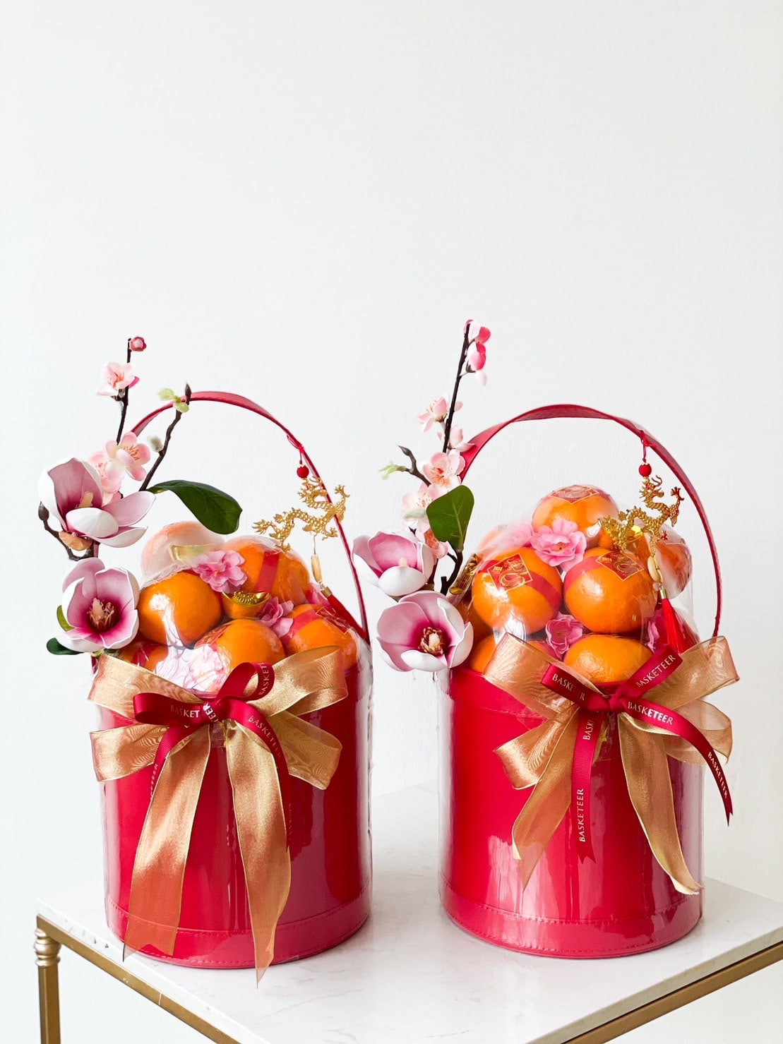 Two red baskets, named the Orange Bliss Box, are filled with oranges and decorated with pink flowers, placed on a white surface. Each basket, perfect for CNY festivities, is adorned with gold and red ribbons tied in bows. Identical in design, they feature handles for easy carrying.