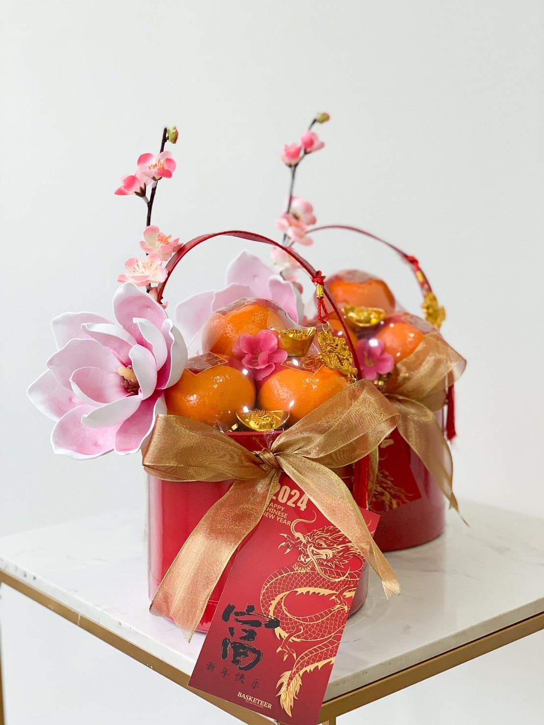 Two red gift baskets filled with oranges are adorned with large pink flowers, branches with smaller pink blossoms, and embellished with gold bows. Each basket has a red tag featuring a dragon illustration and Chinese characters, sitting on a white marble surface.