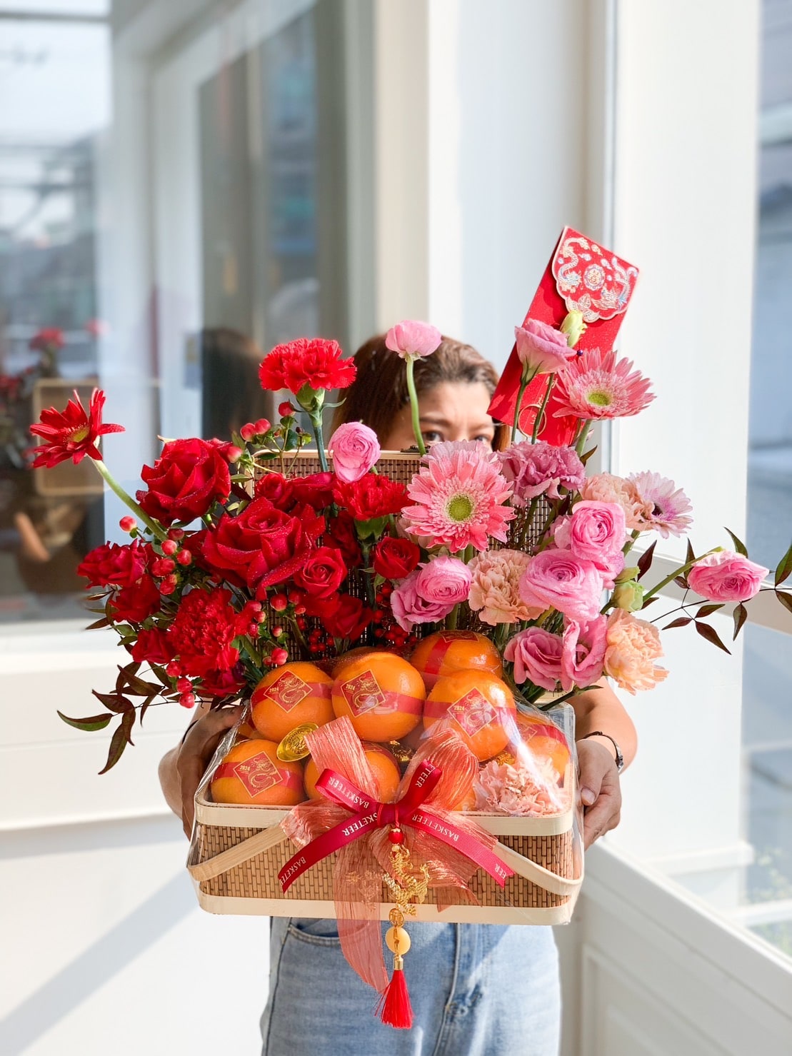 A person holding a rectangular basket adorned with an assortment of red and pink flowers, including roses and carnations, along with several oranges. The basket is decorated with a red ribbon and a red envelope, symbolizing a festive celebration.