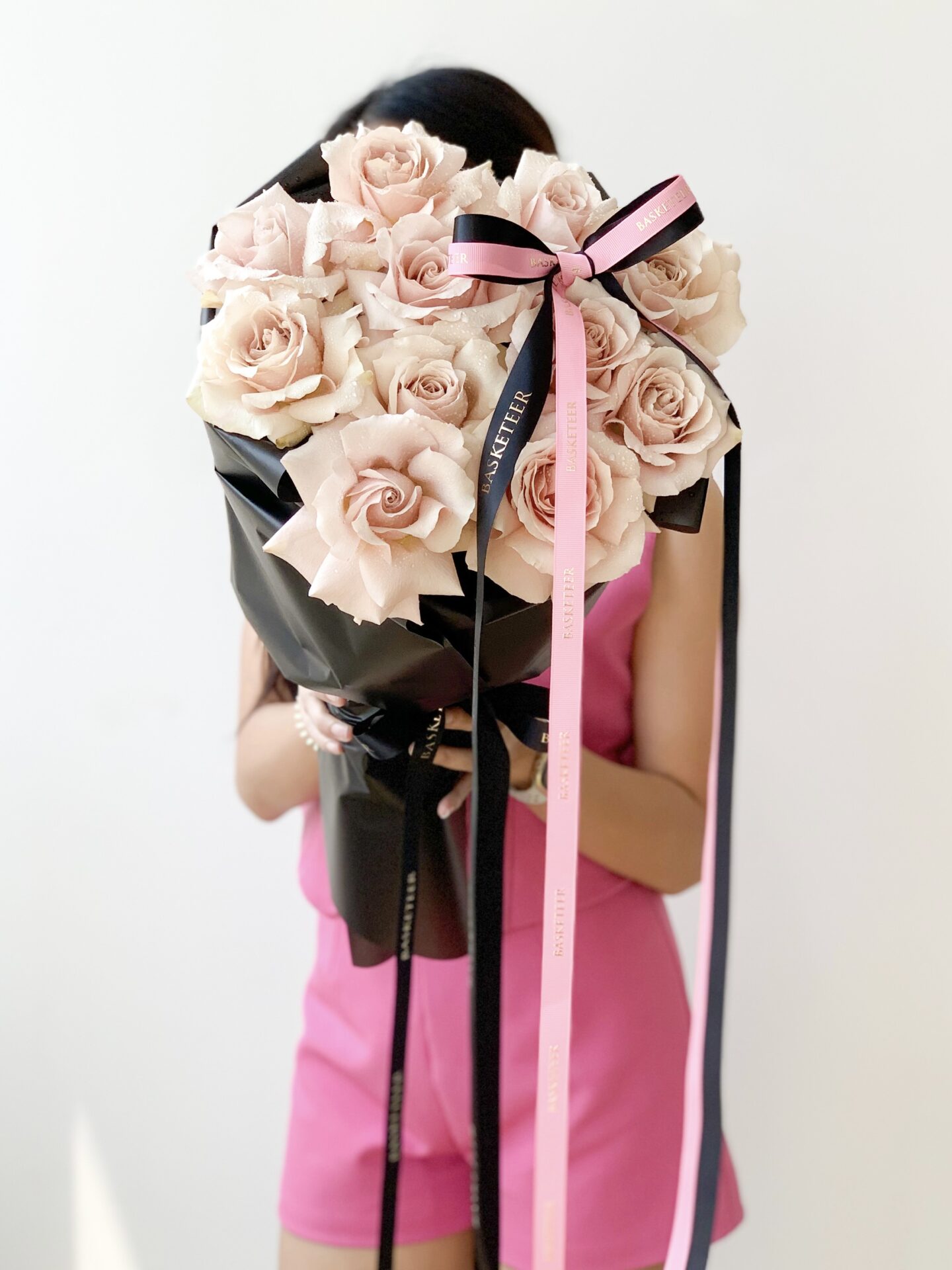 A person in a pink outfit holds a large bouquet of Ocean Song roses wrapped in black paper. The bouquet is adorned with pink and black ribbons. The individual's face is obscured by the flowers, while the background is a plain white wall.
