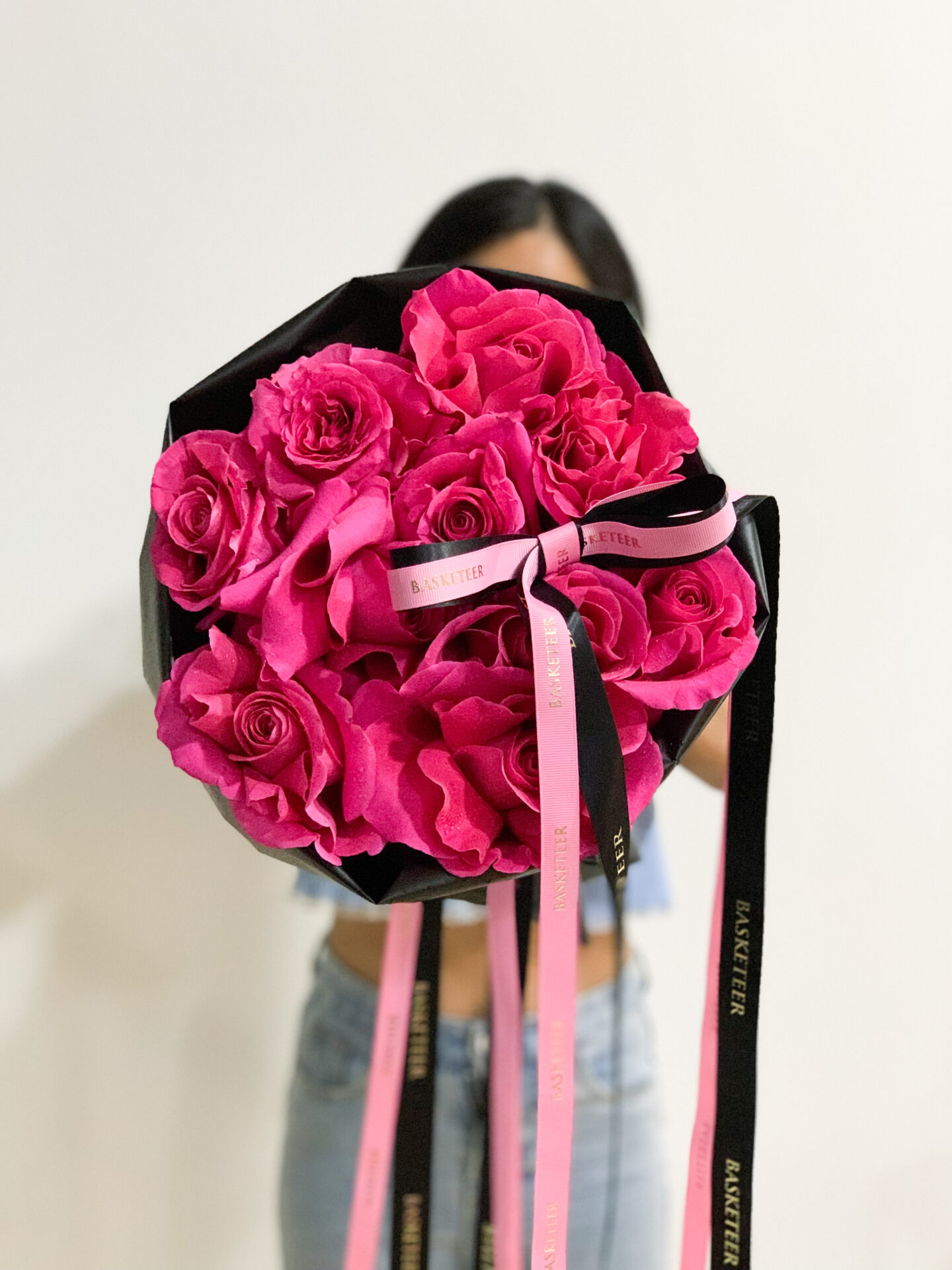 A person is standing and holding a bouquet of vibrant Ocean Song roses wrapped in black paper. Pink and black ribbons are attached to the bouquet, some branded with the word 
