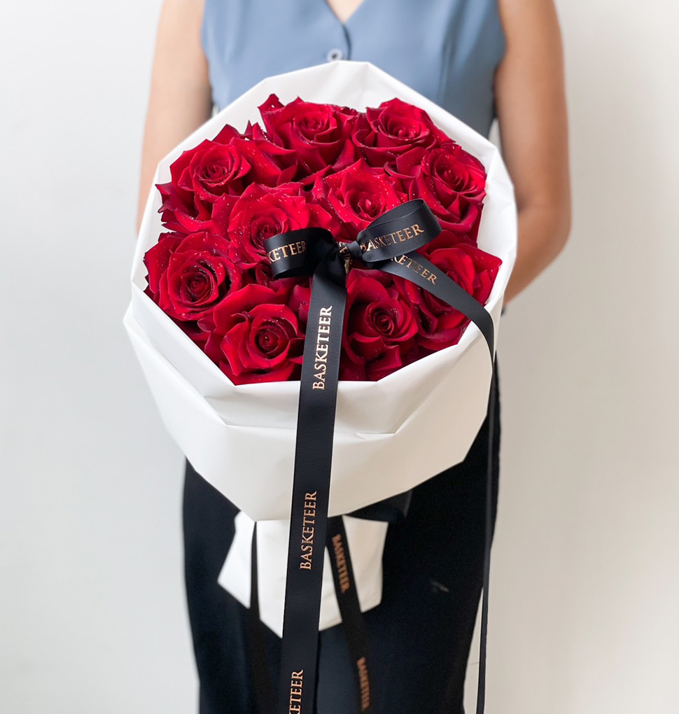 A luxurious hand-tied bouquet of deep red roses, wrapped in elegant white paper with black ribbons, creating a romantic and sophisticated floral arrangement.