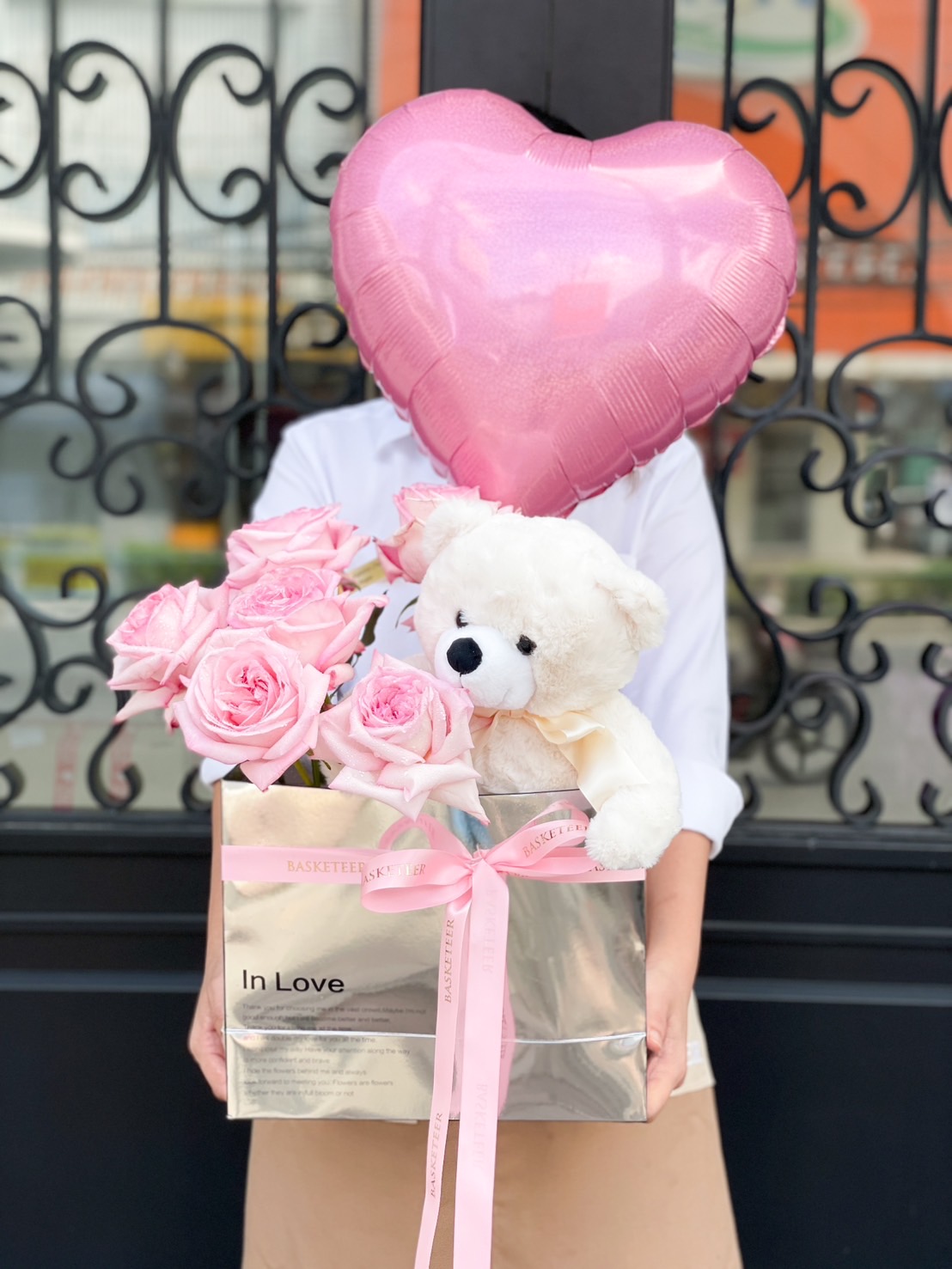 A person holding a bouquet of pink roses and a white teddy bear in a silver gift box wrapped with pink ribbon. A large O'Hara Love Balloon obscures their face, set against the backdrop of a decorative wrought iron gate.