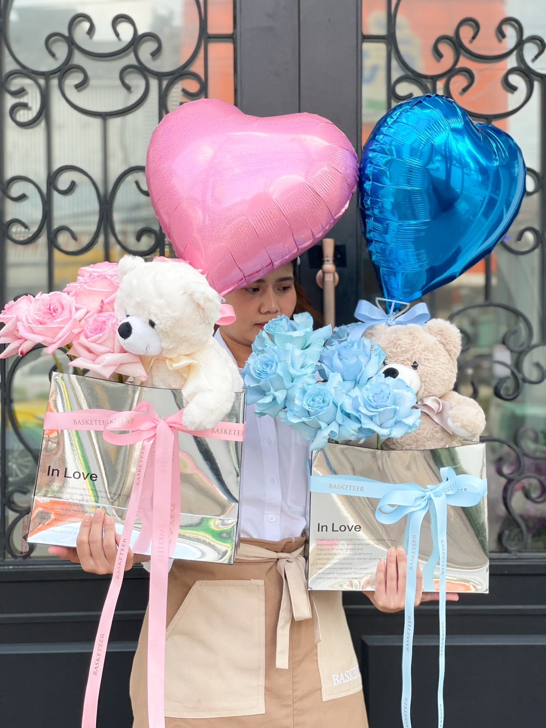 A person standing in front of ornate double doors holds two gift bags. One bag has pink roses, a pink heart balloon, and a white teddy bear. The other bag contains blue roses, a blue heart balloon, and a light brown teddy bear. Both bags, part of the O'Hara Love Balloon Bouquet set, have "In Love" written on them.