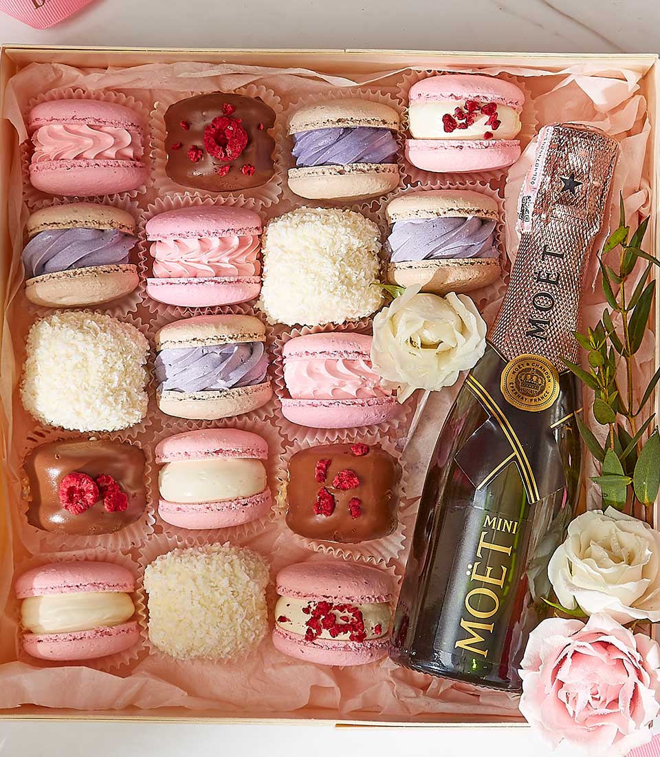 A selection of colorful macarons and desserts arranged in a box with a mini bottle of Moët champagne. The macarons come in various pastel shades, some topped with fruit or drizzled with chocolate. There are also white roses and greenery placed alongside the treats.
