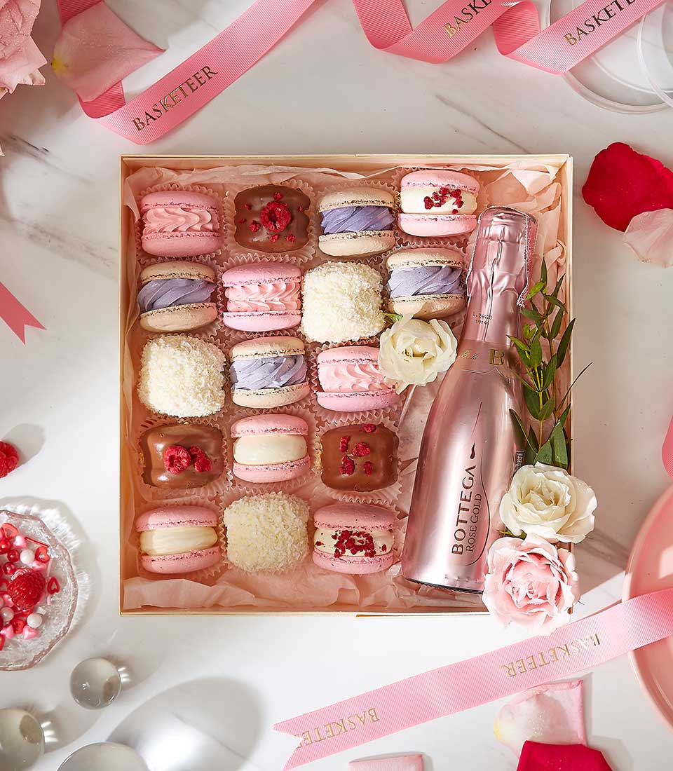 A decorative box containing an assortment of pastel-colored macarons and confections, with a pink bottle of sparkling wine. The box is adorned with pink and white roses and greenery. The scene includes scattered roses, ribbons, and other decorative elements.