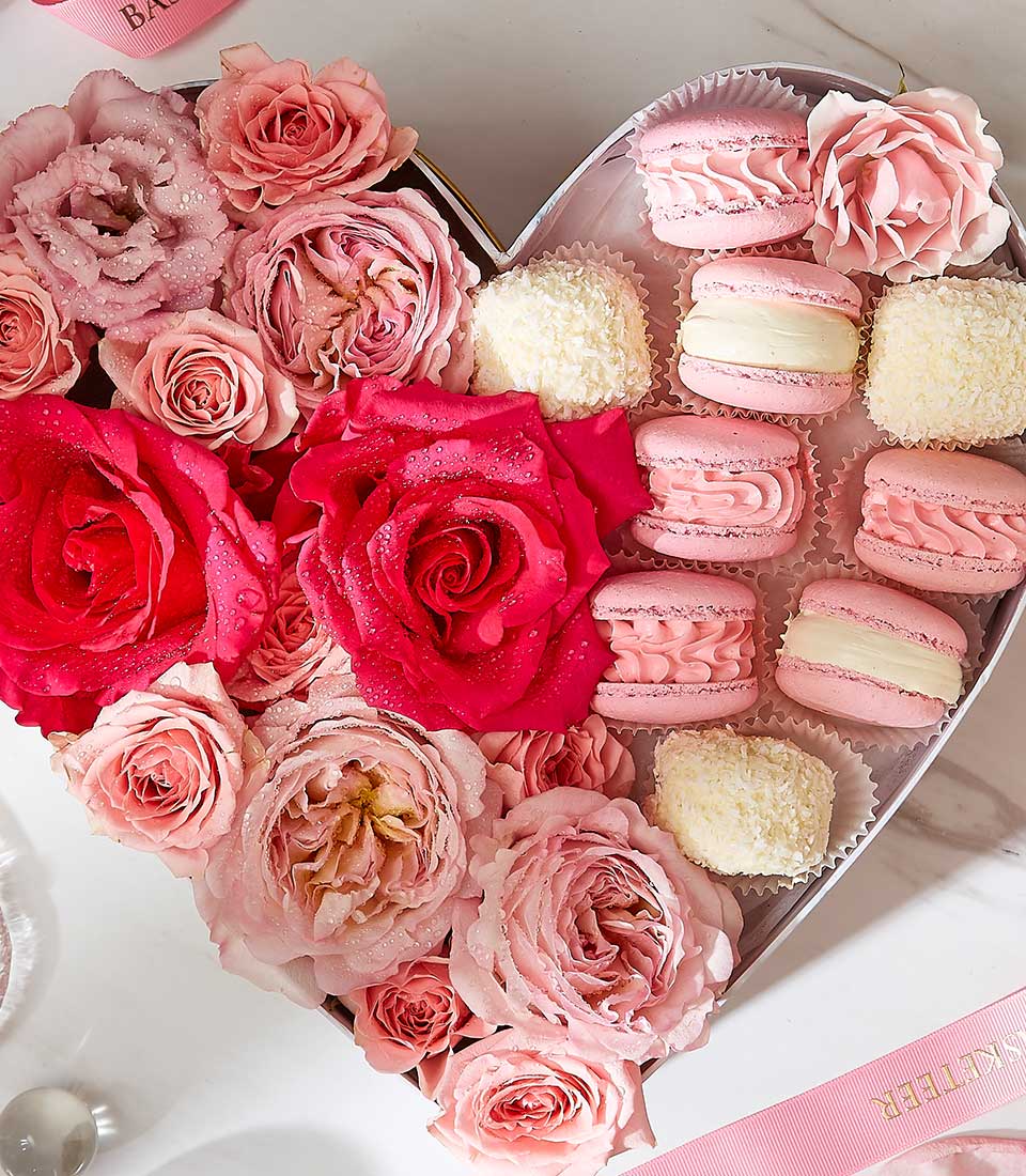 A heart-shaped box containing an arrangement of pink roses alongside an assortment of sweet macarons and pastries. The box sits on a white marble surface with a pink ribbon visible at the edge.