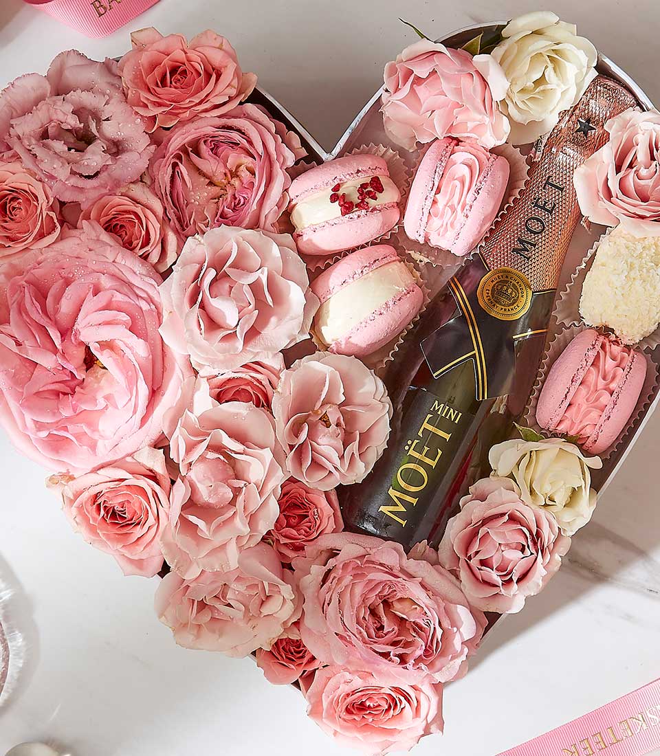 A heart-shaped arrangement featuring a bottle of Moët champagne, surrounded by pink and white roses, and several macarons and sweets. The overall presentation is romantic and luxurious, perfect for special occasions or celebrations.