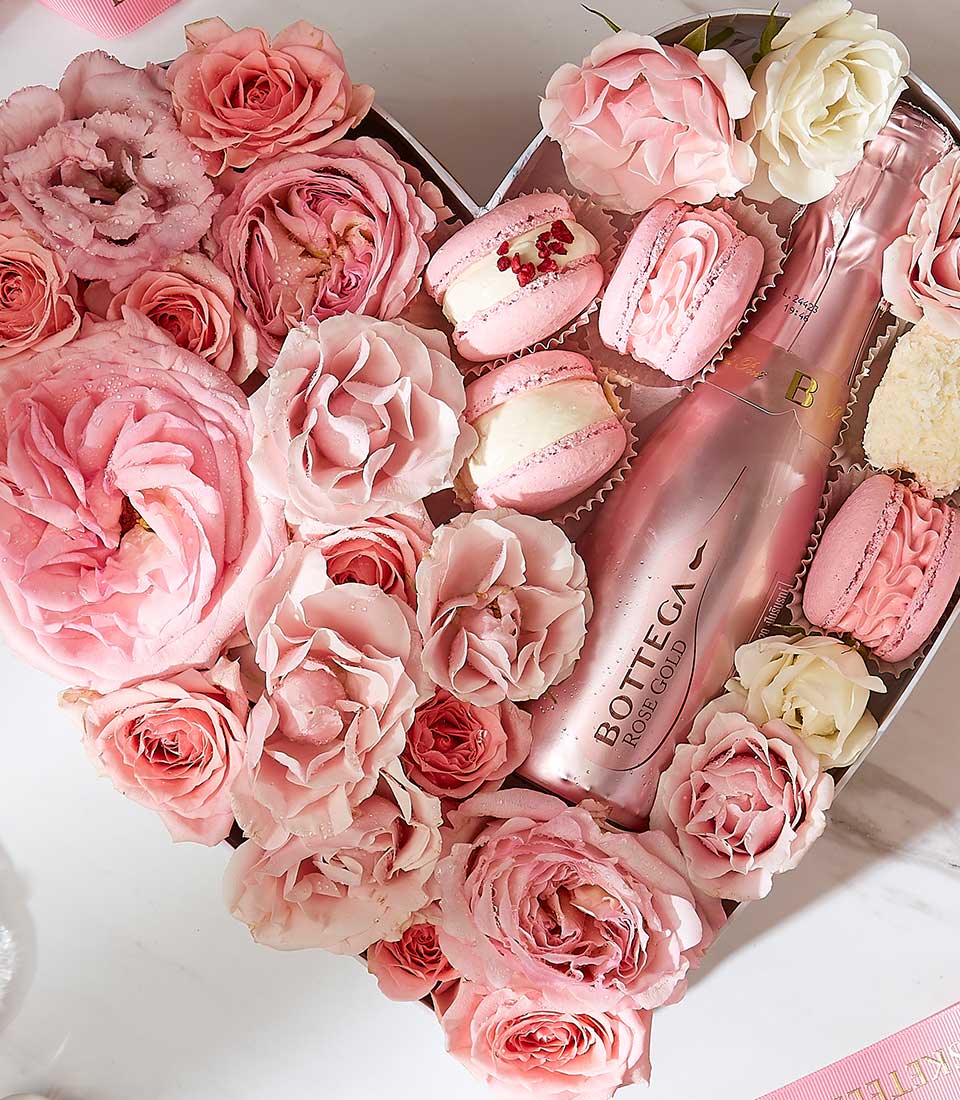 A pink-themed gift box is filled with a bottle of Bottega Rose Gold sparkling wine, pink and white roses, and assorted pink macarons. The bottle is nestled among the flowers and treats, creating a luxurious and elegant presentation.
