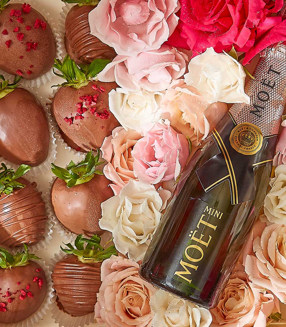 A bottle of Moët Mini champagne lies surrounded by various pink, white, and red roses. Several chocolate-coated strawberries, some decorated with sprinkles, are arranged nearby in white paper cups, creating an elegant and romantic presentation.