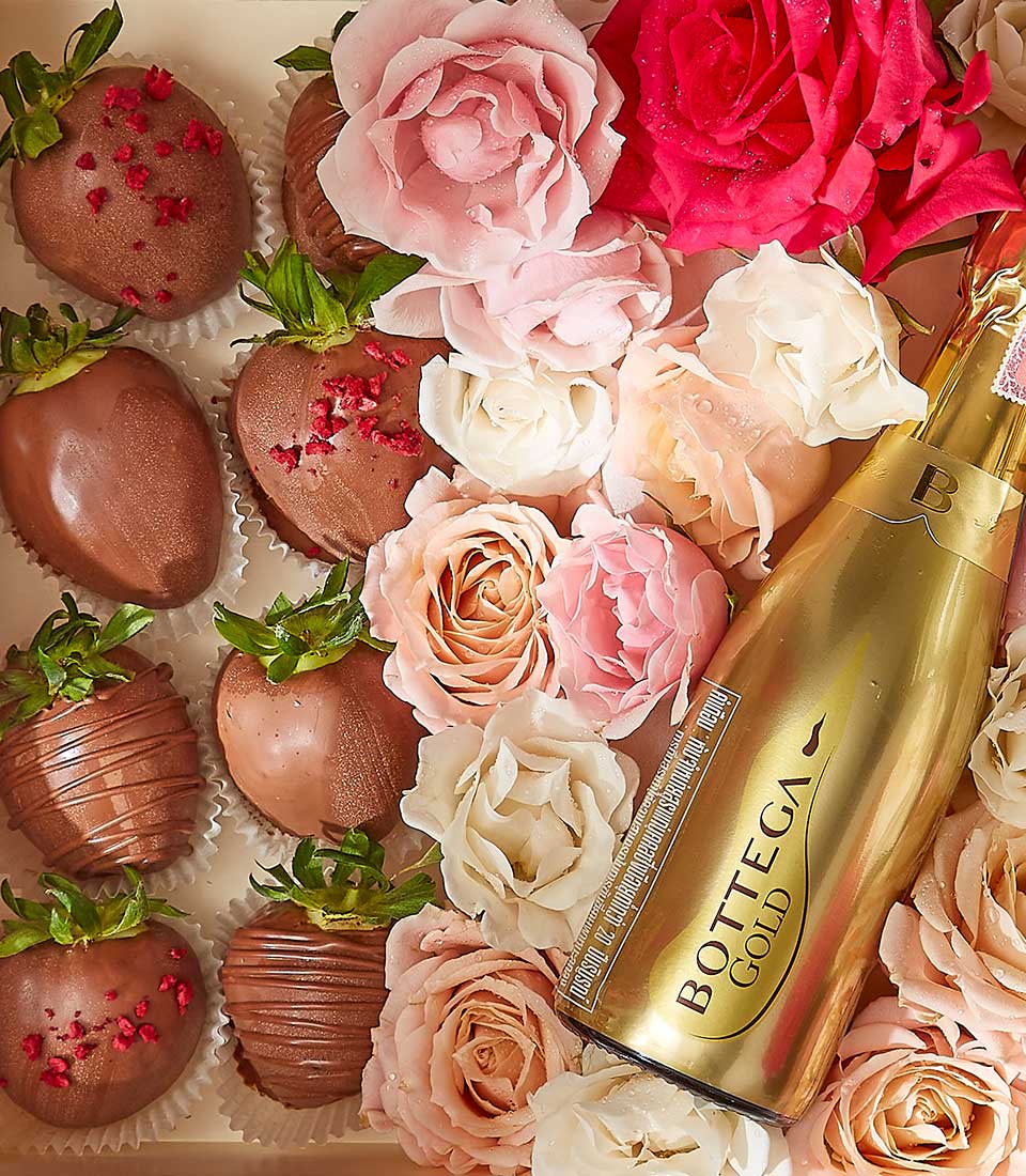 A luxurious arrangement featuring a gold bottle of Bottega Gold prosecco, surrounded by pink and white roses and chocolate-covered strawberries, some adorned with red sprinkles, all beautifully presented in an elegant gift box.