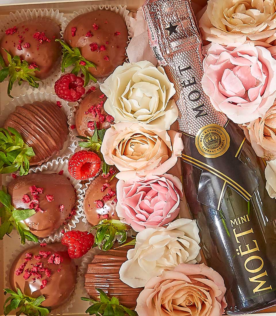 A gift box containing a bottle of Moët mini champagne, chocolate-covered strawberries, fresh raspberries, and an assortment of pink, white, and peach roses. The Chocolate-Dipped Strawberry Wine blooms with strawberries decorated in chocolate drizzle and sprinkled with freeze-dried raspberries.