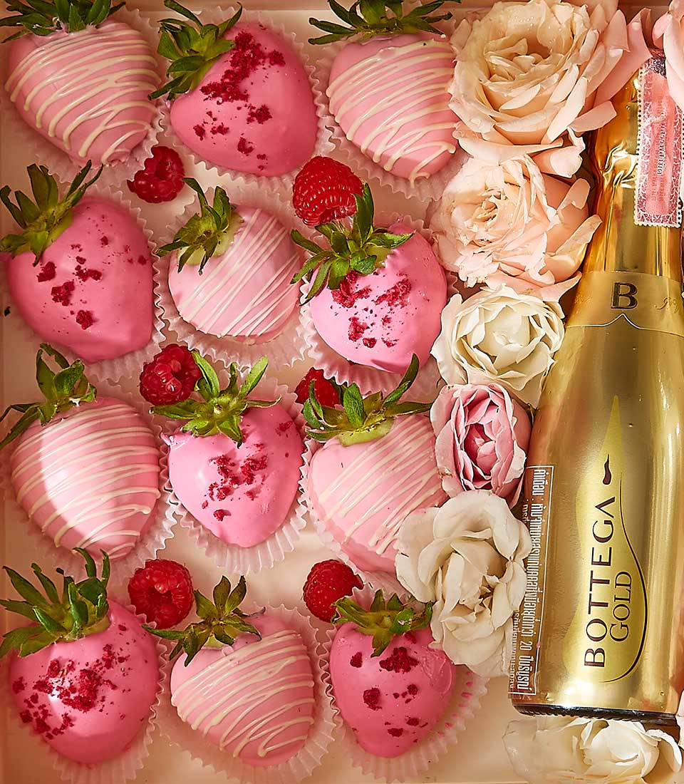 A beautifully arranged box containing pink chocolate-covered strawberries, fresh raspberries, and an assortment of white and pink flowers. A bottle of Bottega Gold Prosecco is also included on the right side, adding an elegant touch to the presentation.