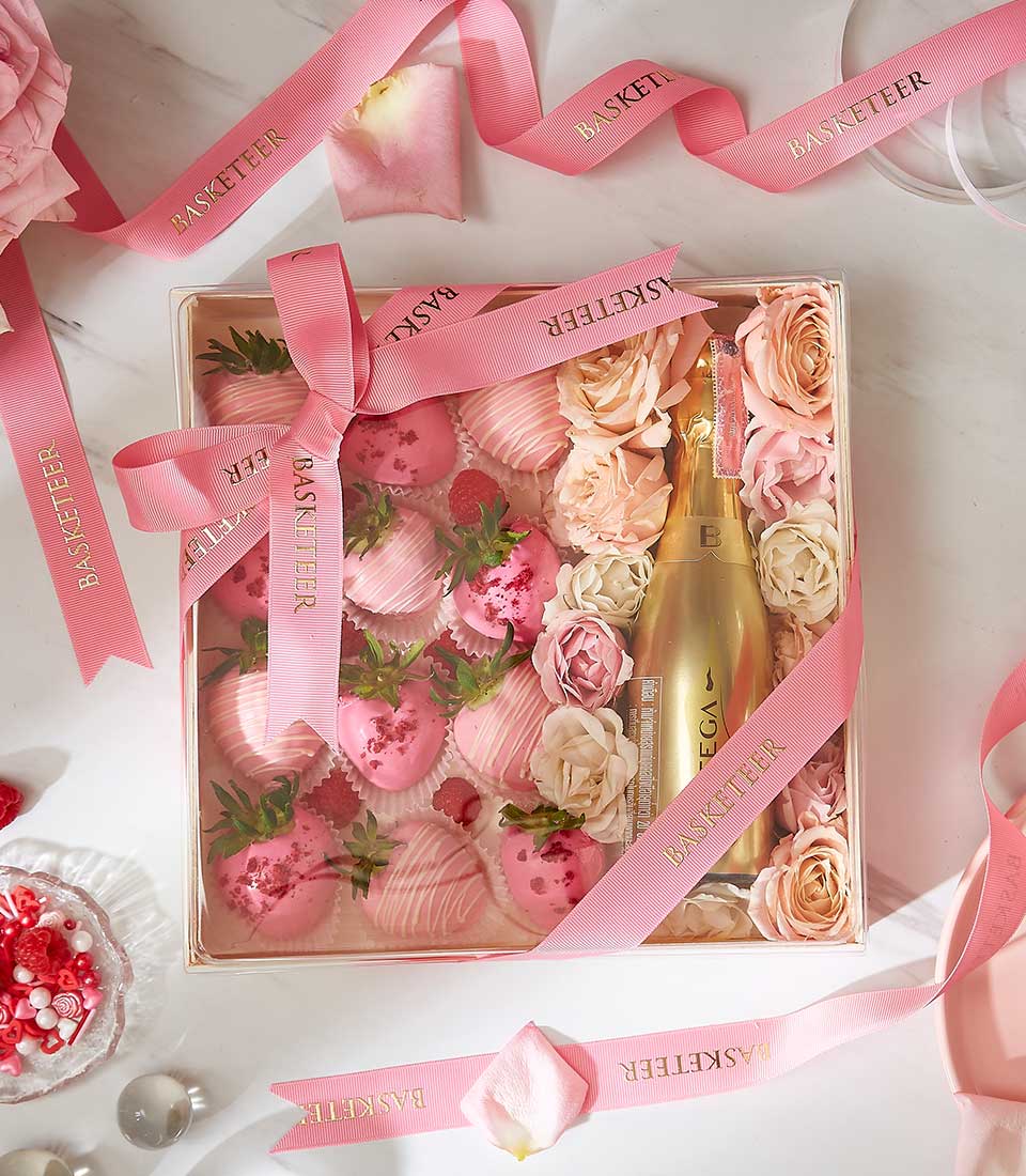 A gift box featuring pink chocolate-dipped strawberries, a bottle of champagne, and an assortment of roses. The box is adorned with pink ribbons labeled 