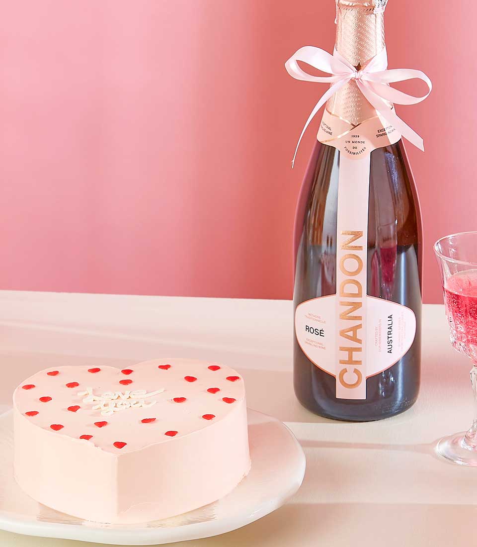 A heartfelt wine cake gift set features a heart-shaped cake with pink frosting and red polka dots, sitting on a white plate beside a bottle of Chandon Rosé sparkling wine adorned with a white ribbon. A half-full glass of pink bubbly sits next to the bottle, all set against a plain pink background.