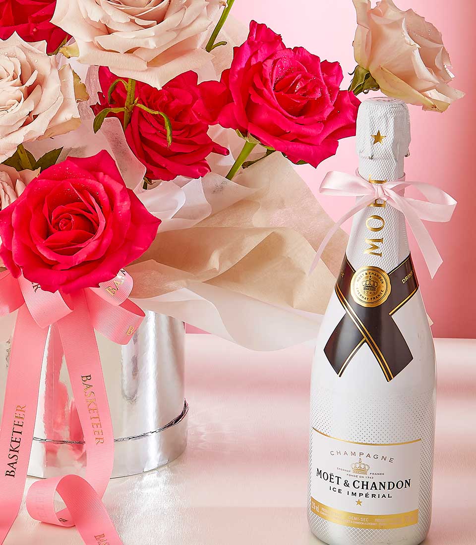 A bouquet of red and pink roses in a white vase adorned with pink ribbons beside a white bottle of Moet & Chandon Ice Imperial champagne. The bottle has gold accents and a white ribbon tied around its neck. The background features a gradient from pink to white.