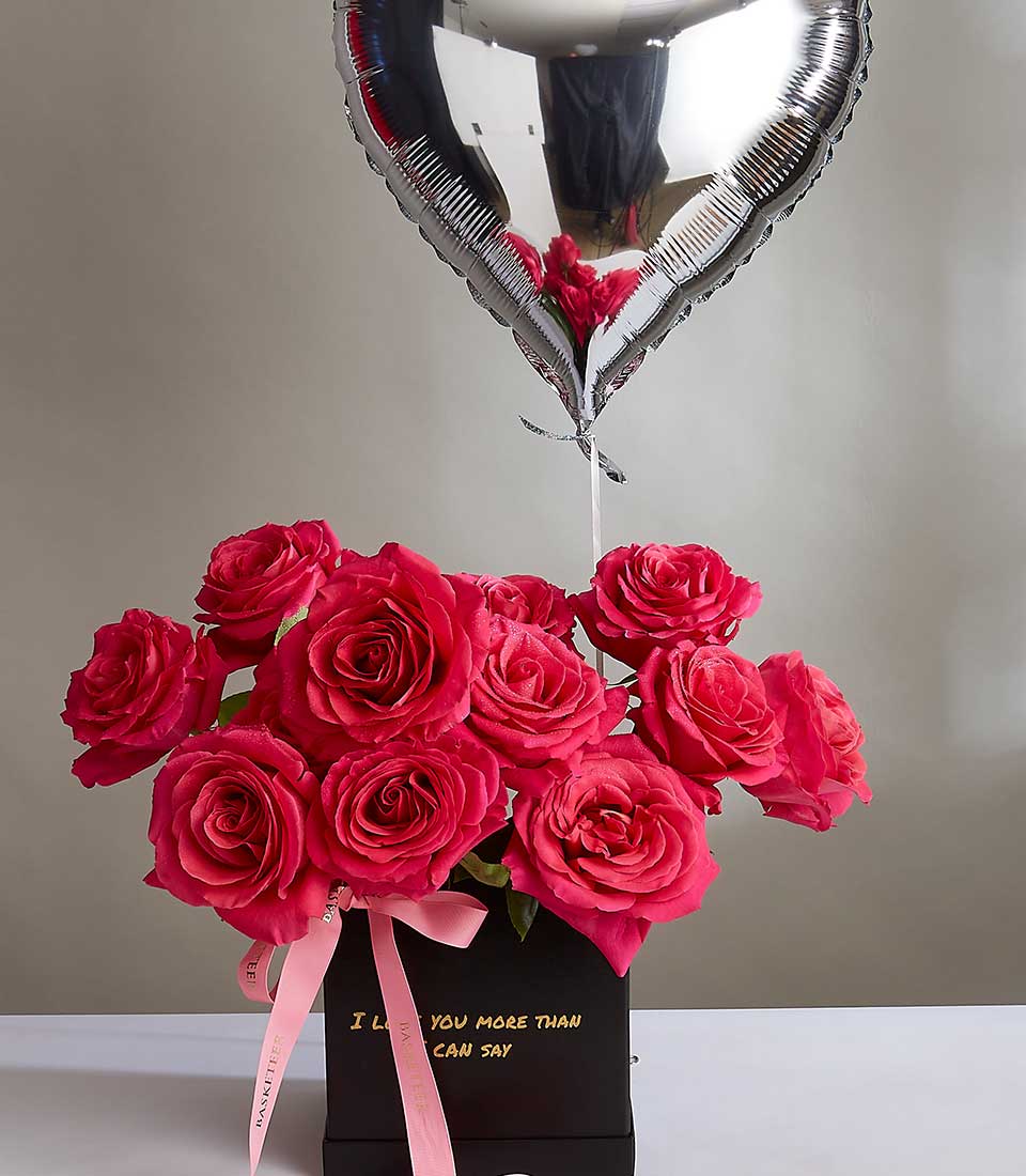A bouquet of vibrant red roses in a black rectangular vase with a pink ribbon tied around the stems. The vase, reminiscent of Pink Floyd's iconic imagery, bears a heartfelt message. A silver heart-shaped balloon floats above the bouquet, reflecting light in its metallic surface.